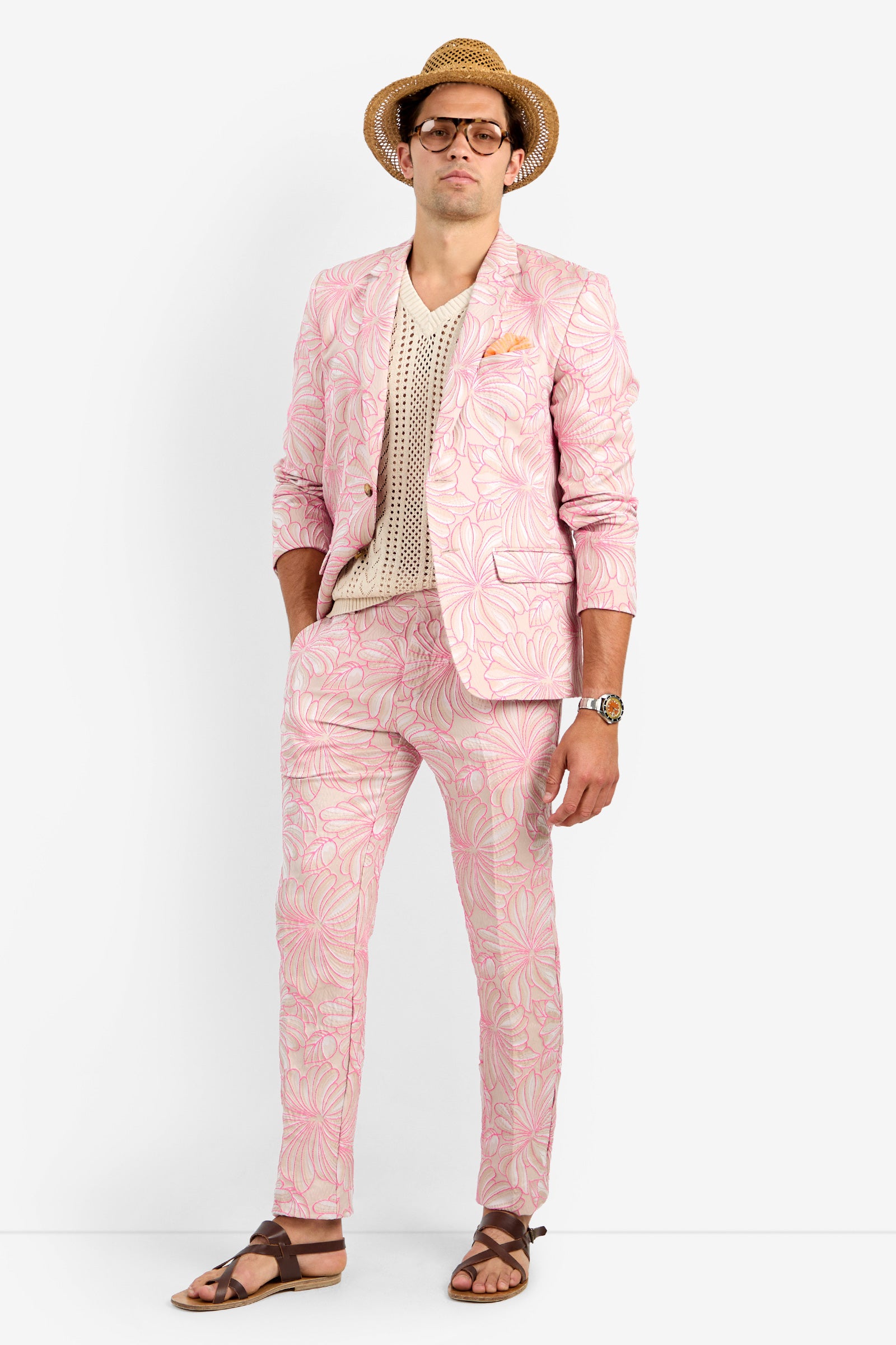 A man stands against a plain background wearing the THURSTON BLAZER in pink, paired with a cream crochet shirt, brown sandals, a straw hat, and glasses. His hands are in his pockets and he has a neutral expression.