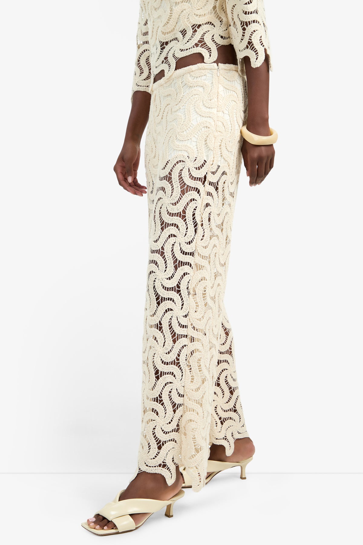 A person wearing the BARDAN PANT in cream, with a matching crochet-patterned cotton top, cream heeled sandals, and a chunky bangle stands against a plain white background.