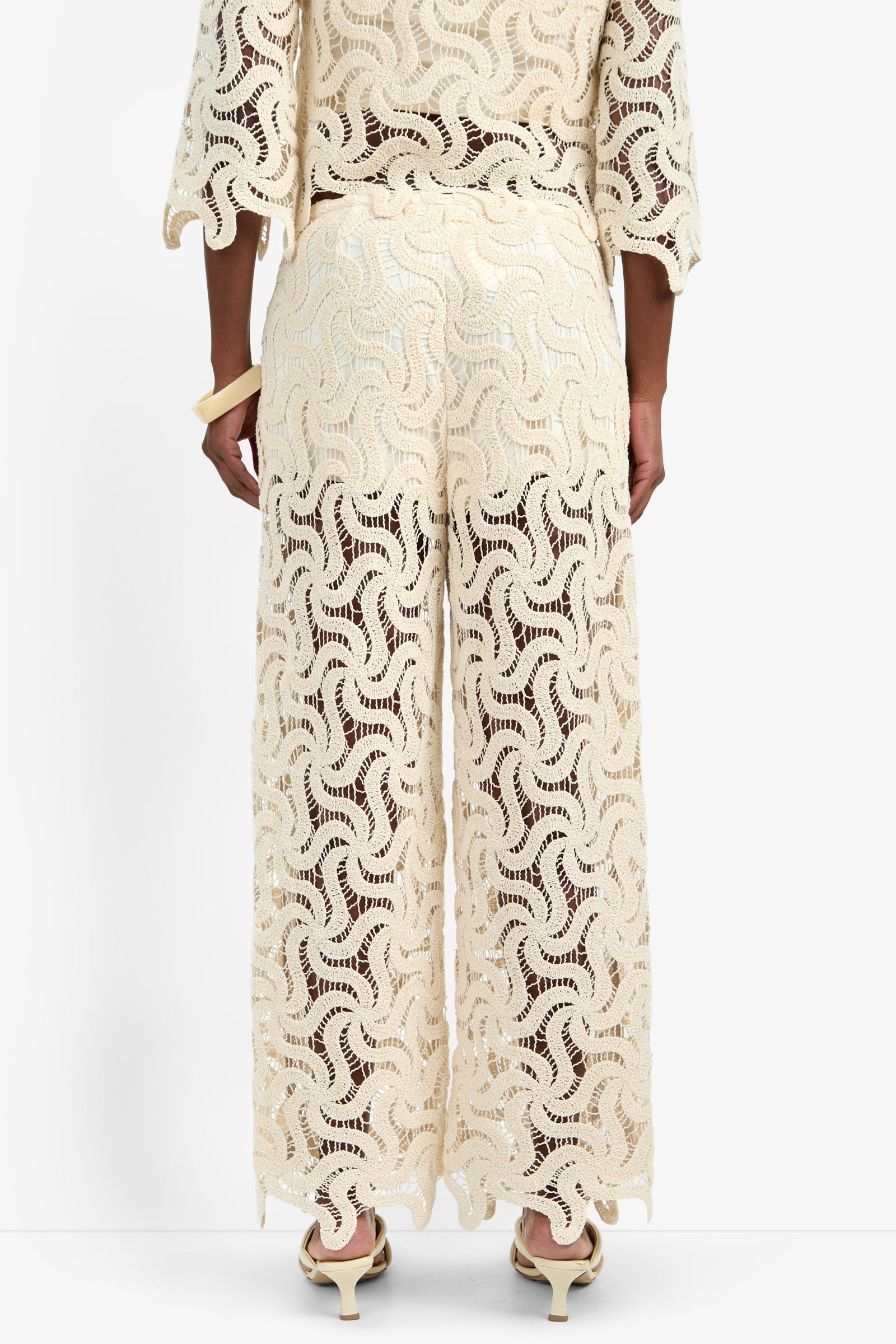 A person stands facing away, wearing the BARDAN PANT—cream-colored, wide-leg cotton pants with a wavy lace pattern. Semi-sheer and stylish, they're paired with a matching top and cream high-heeled sandals.