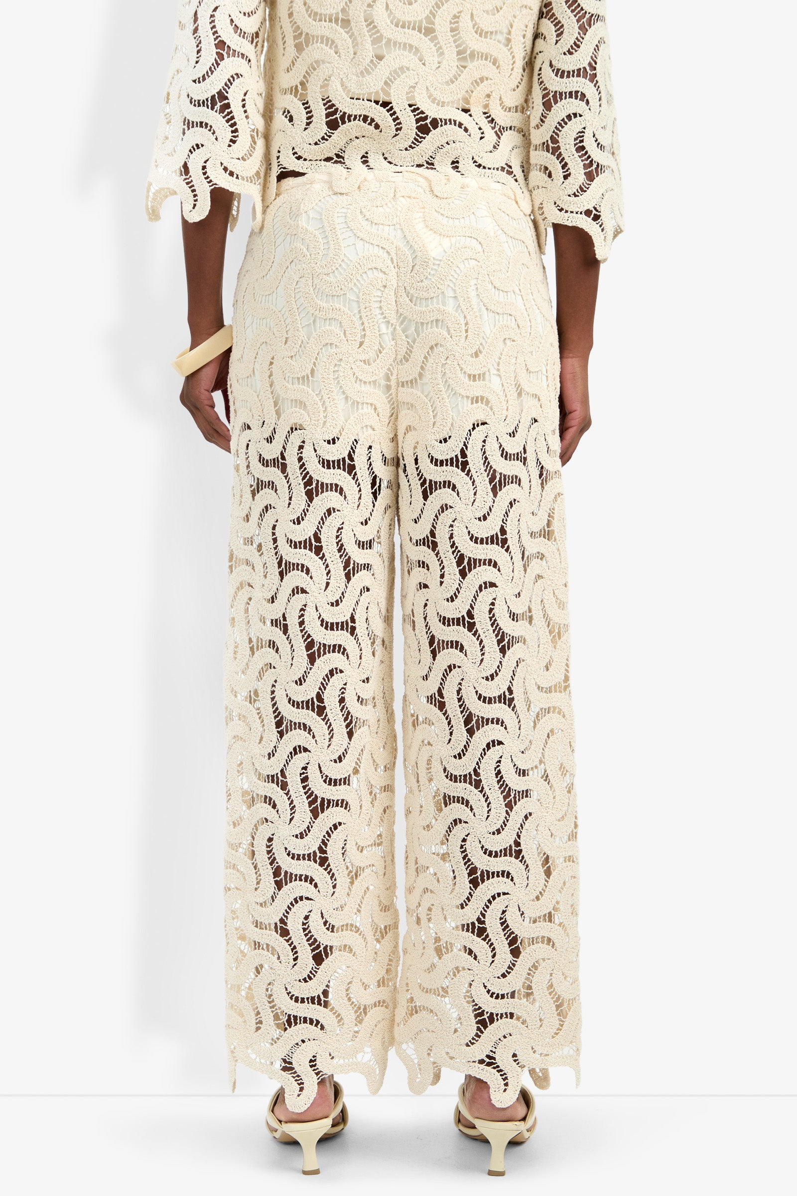 A person faces away, wearing the cream Bardan Pant with wide legs and intricate crochet lace. Paired with a matching top, they sport light shoes and a large bracelet against a plain white background.