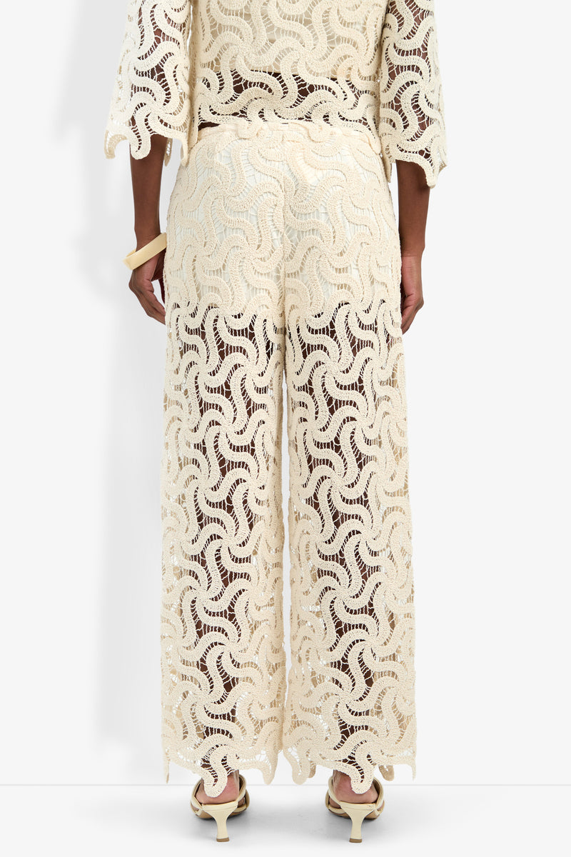 A person faces away, wearing the cream Bardan Pant with wide legs and intricate crochet lace. Paired with a matching top, they sport light shoes and a large bracelet against a plain white background.