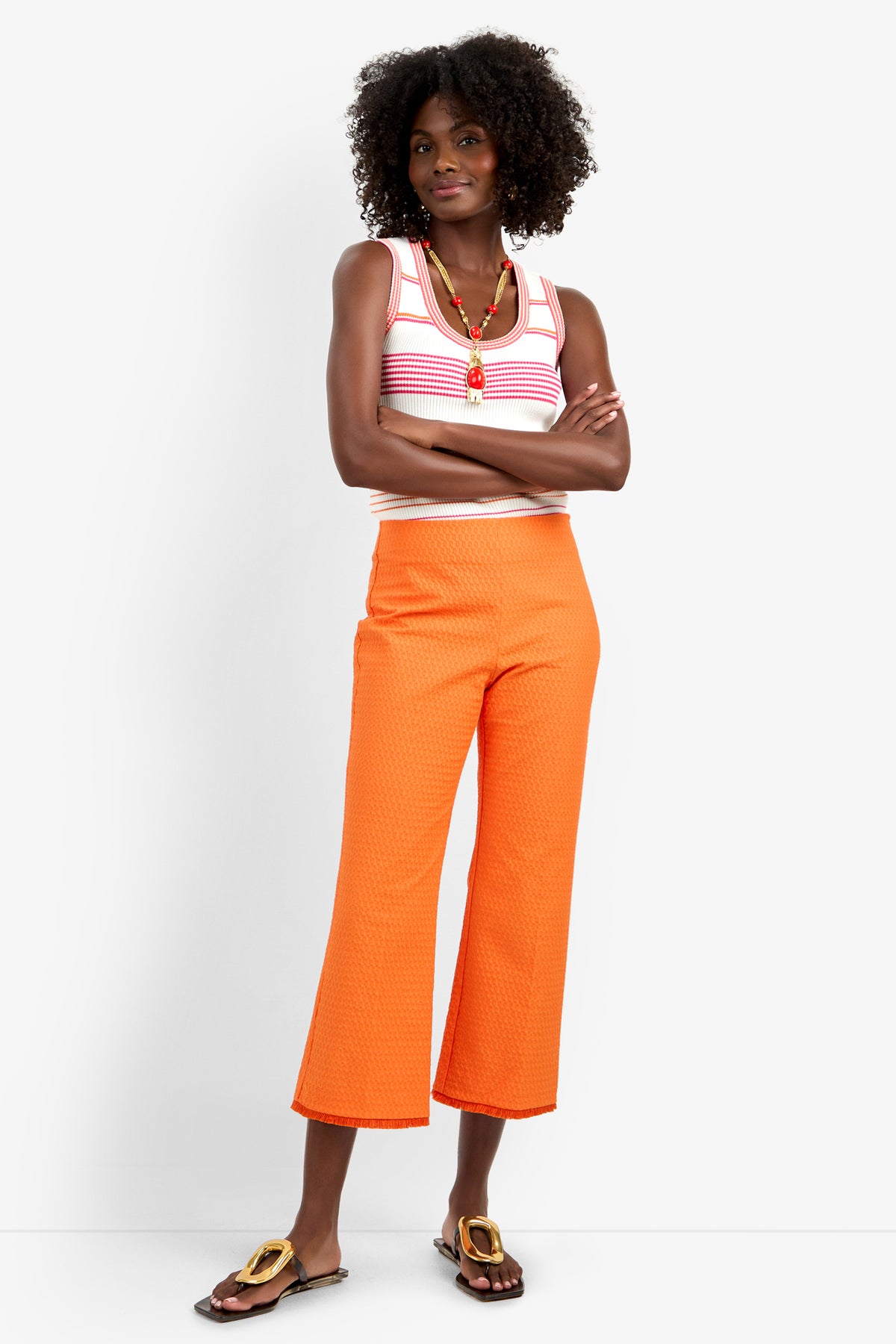 A woman with curly hair poses against a plain white background, wearing the GULF PANT in orange, a sleeveless white and pink striped viscose top, a colorful necklace, and brown sandals. Her arms are crossed.