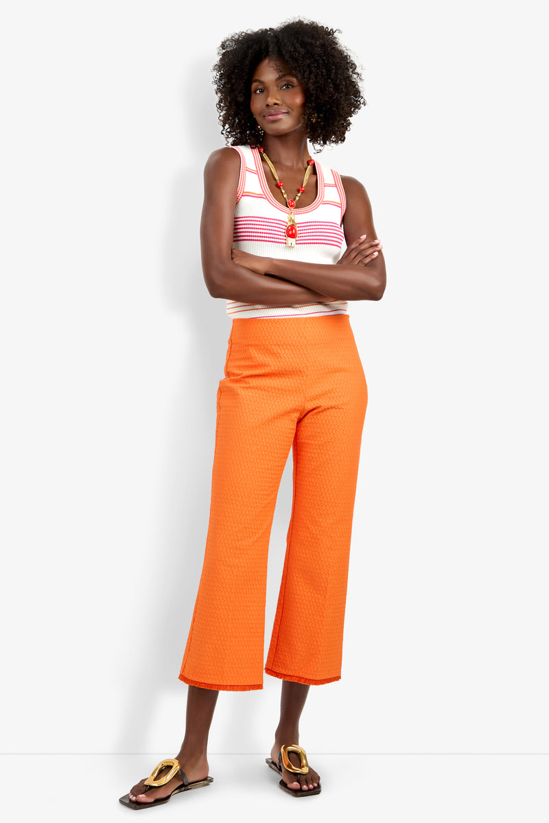 A woman with curly hair and a bright smile stands against a white background, arms crossed, wearing the Gulf Pant in orange, a sleeveless striped tank top, sandals, and a colorful beaded necklace.