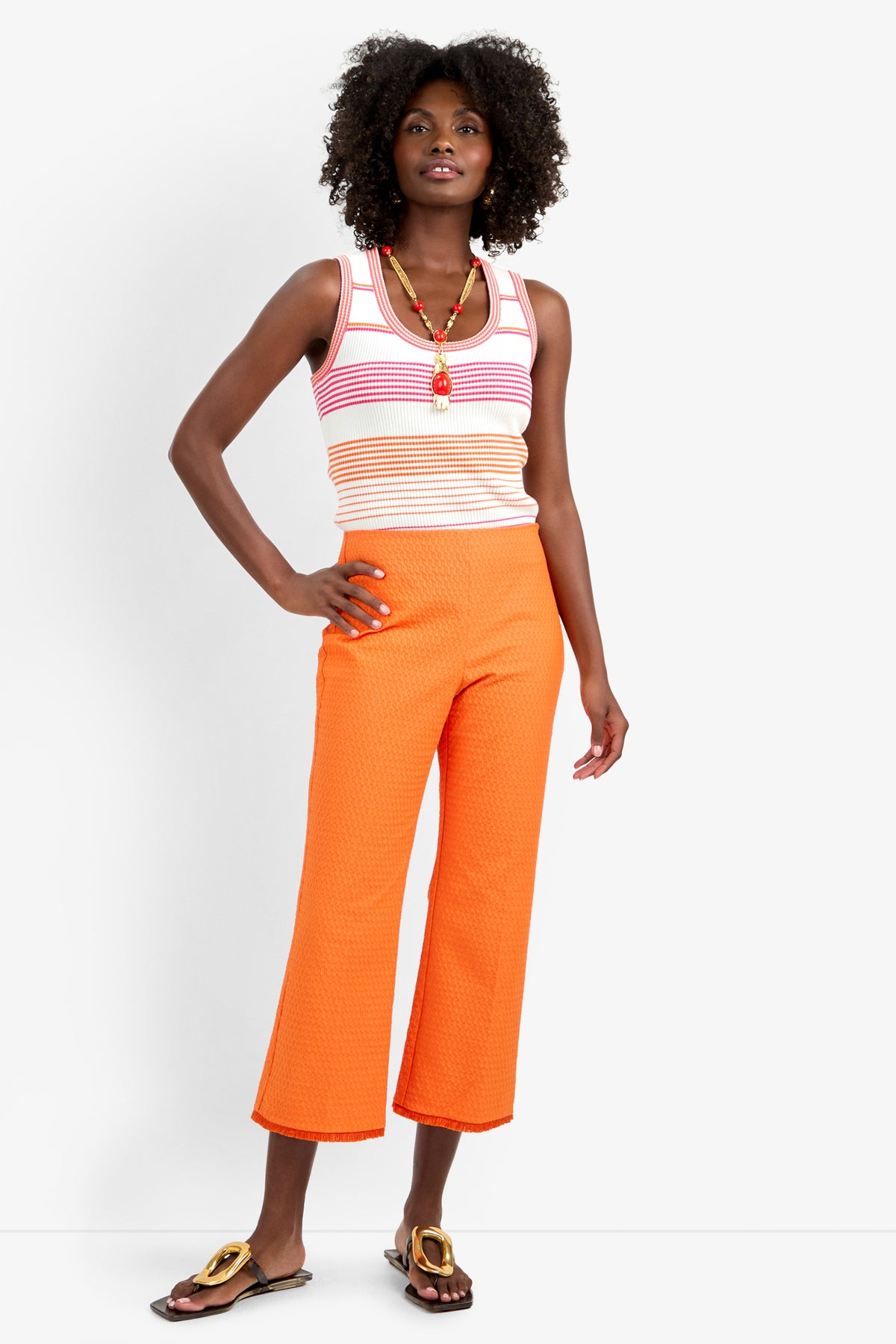 A woman with curly hair poses confidently against a white background, wearing the GULF PANT—orange textured wide-leg pants—paired with a sleeveless pink and white striped viscose tank, gold sandals, and layered necklaces.