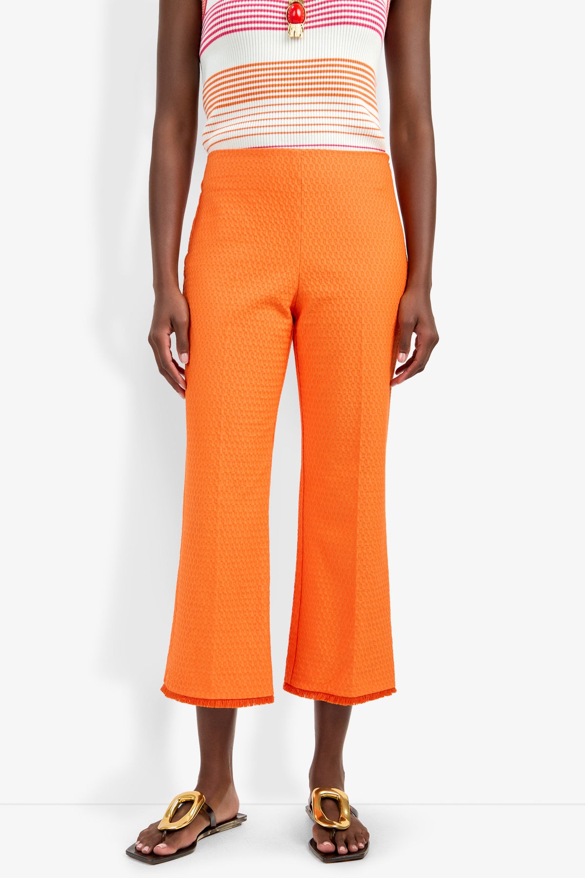 A person wearing Gulf Pant in bright orange with a textured, cropped design, paired with a colorful striped sleeveless top and brown sandals featuring gold embellishments, stands against a white background.
