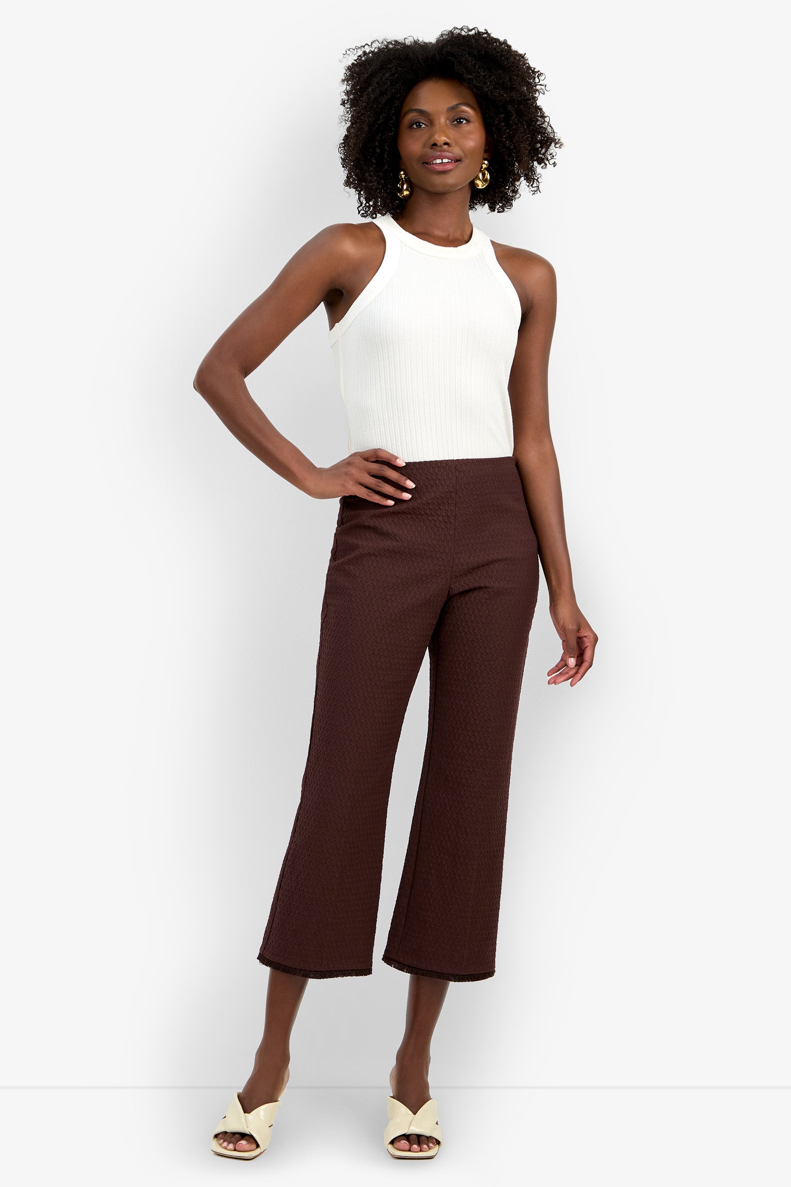 Woman wearing a sleeveless white ribbed top, the high-waisted Gulf Pant in brown, cream open-toe heels, and yellow earrings, standing against a plain white background.
