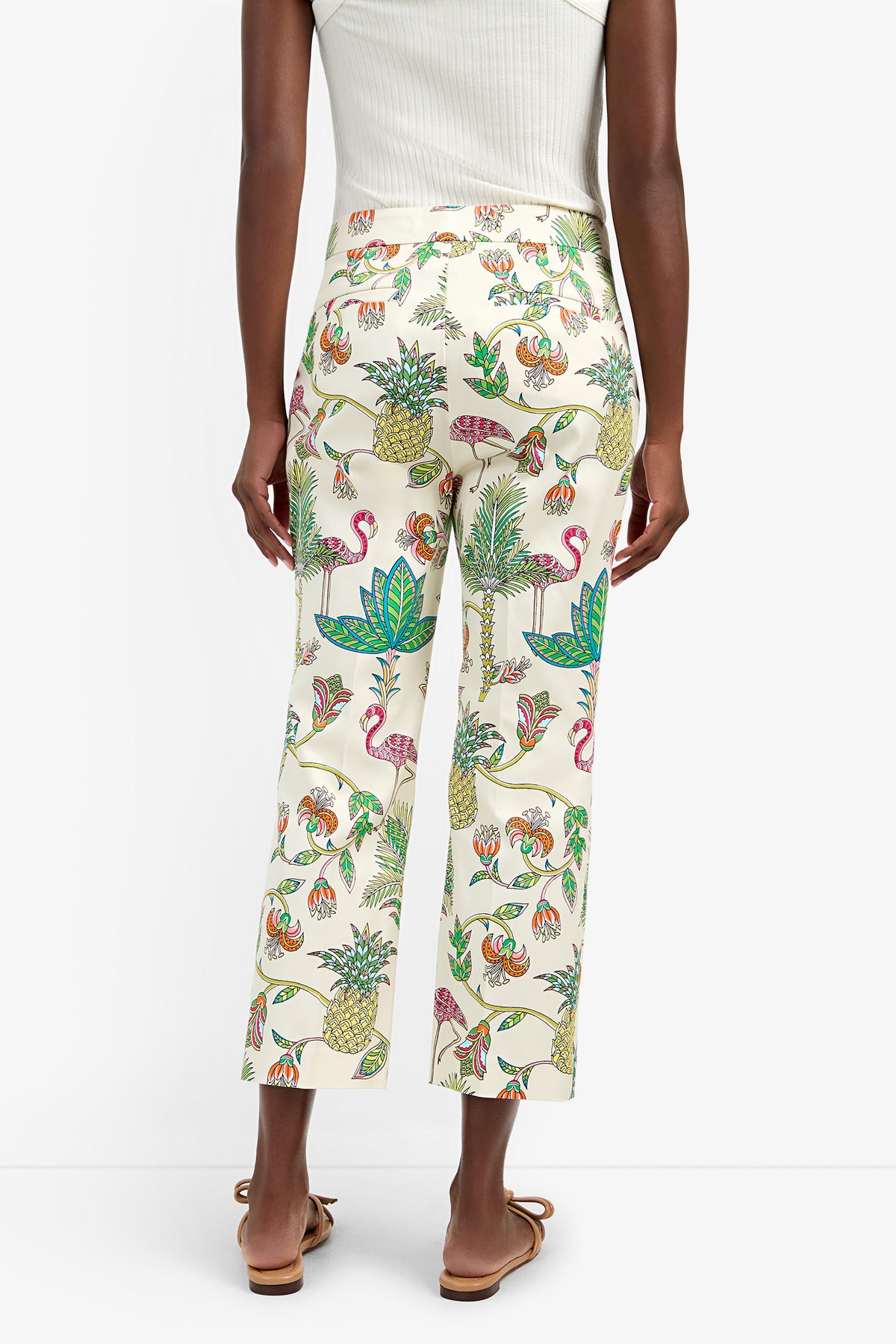 A person is seen from the back wearing the LULU PANT—cream-colored cotton pants with vibrant tropical prints (flamingos, pineapples, leaves)—paired with a white sleeveless top and flat sandals, set against a plain background.