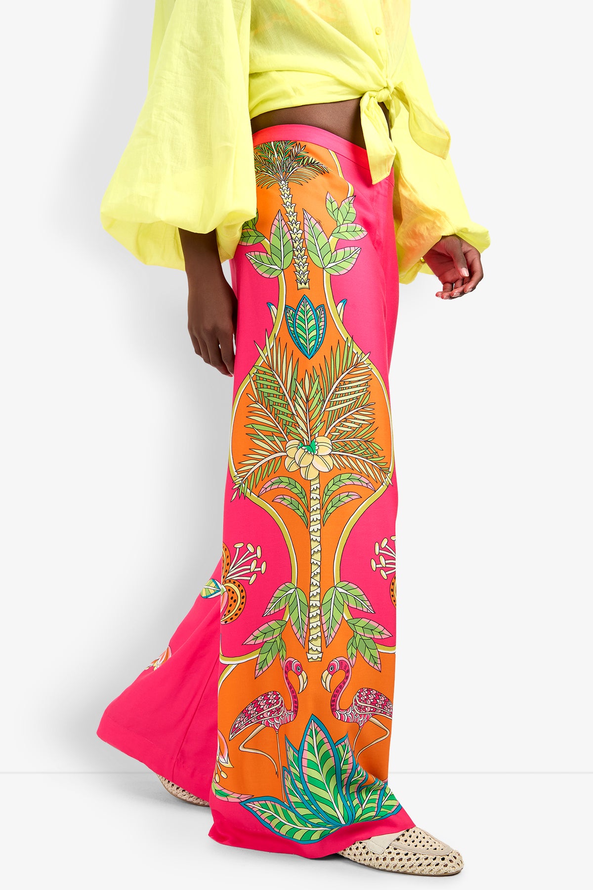 Someone wearing the Lanay Pant, featuring a vibrant tropical print in pink, orange, and green with a wide-leg fit, paired with a light yellow tie-front shirt and beige woven pointed shoes.