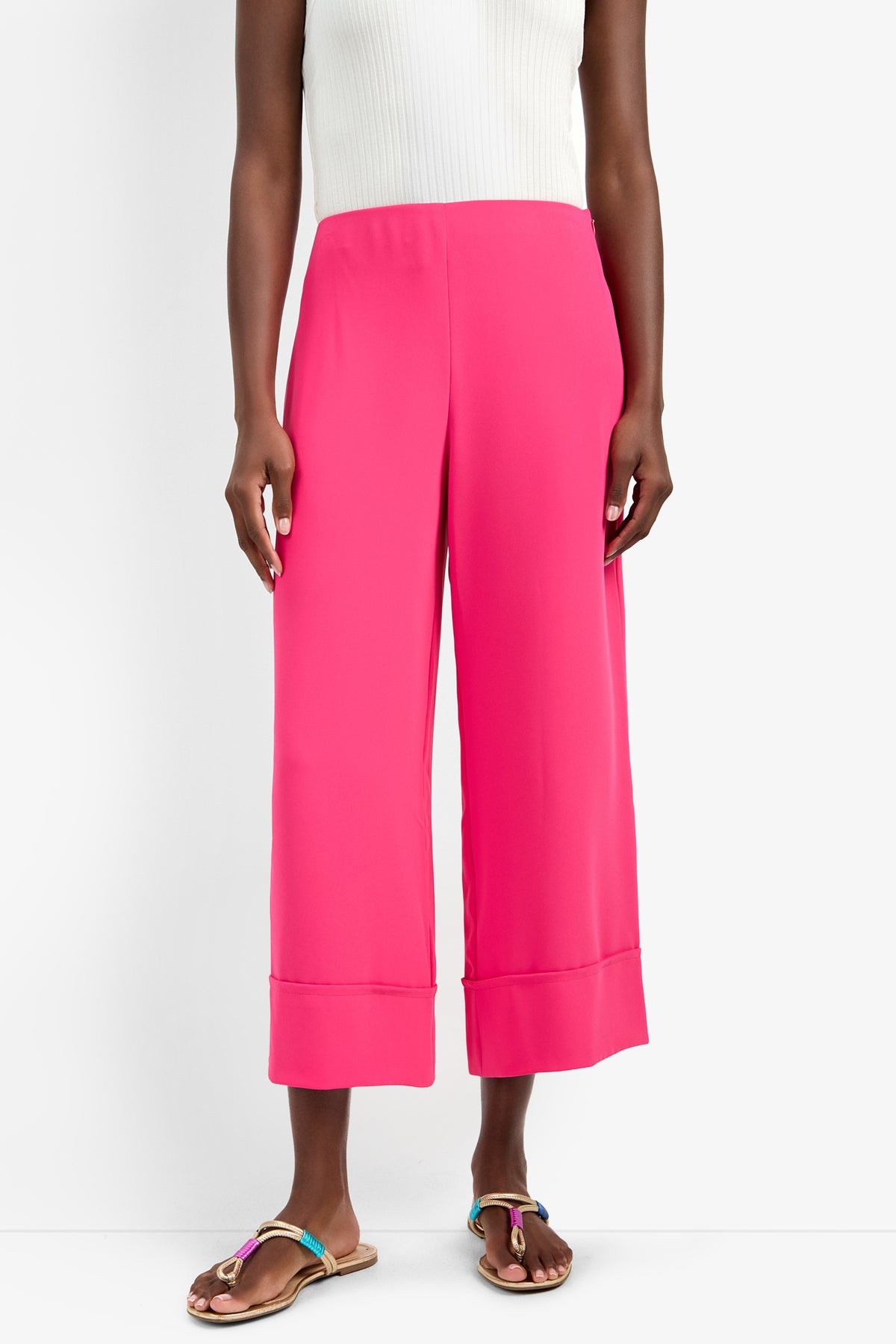 Wearing bright pink BAHOOKA PANT wide-leg cropped pants (100% polyester), a white ribbed sleeveless top, and brown flat sandals with colorful straps, the person stands against a white background.