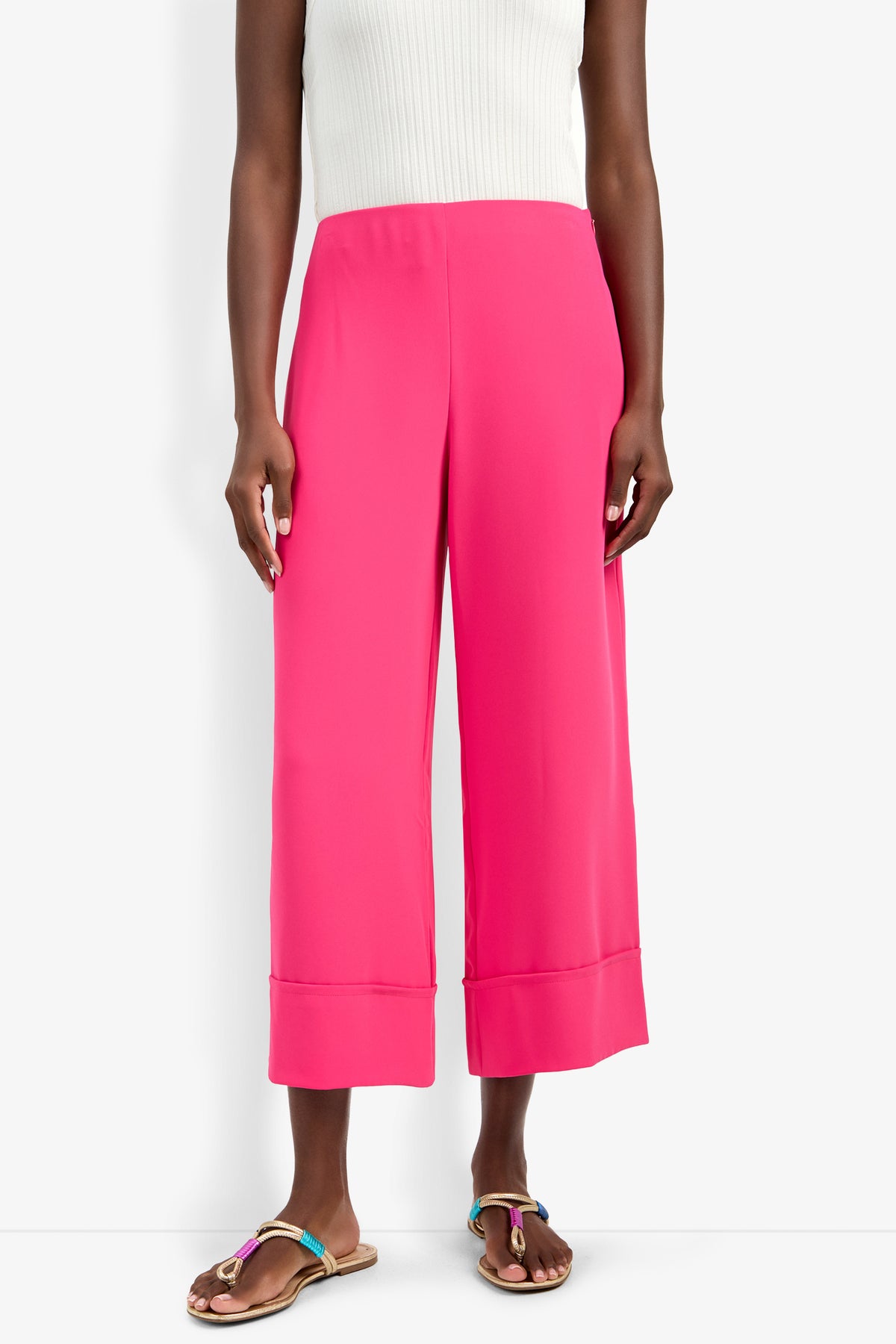 A person wearing the Bahooka Pant in bright pink, styled with a white sleeveless ribbed top and tan flat sandals with colorful straps, stands against a white background.