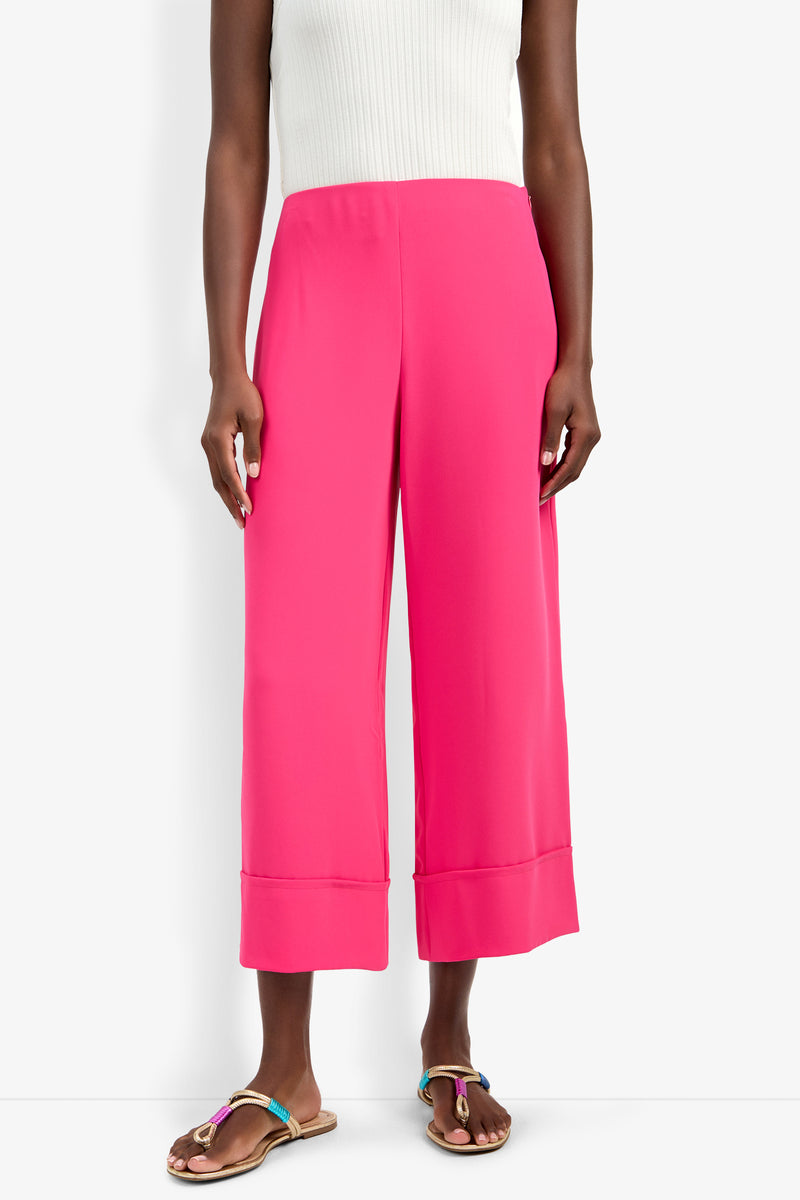A person wearing the Bahooka Pant in bright pink, styled with a white sleeveless ribbed top and tan flat sandals with colorful straps, stands against a white background.