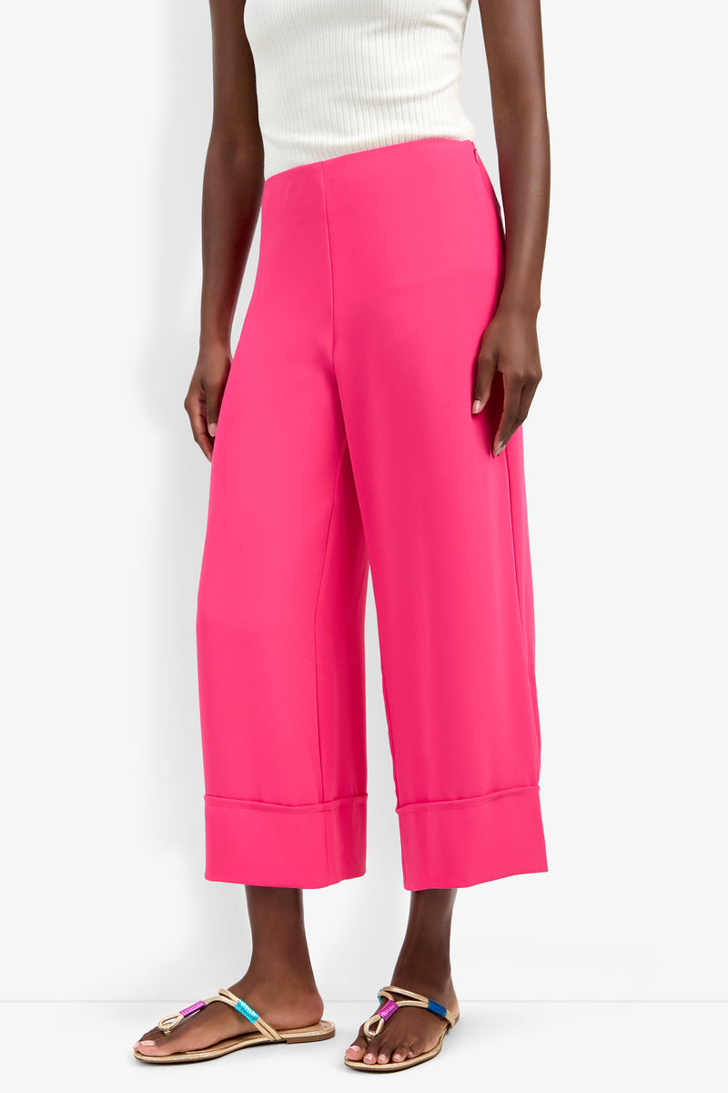A person wearing the Bahooka Pant in bright pink, paired with a white sleeveless ribbed top and colorful flat sandals, stands against a plain white background.
