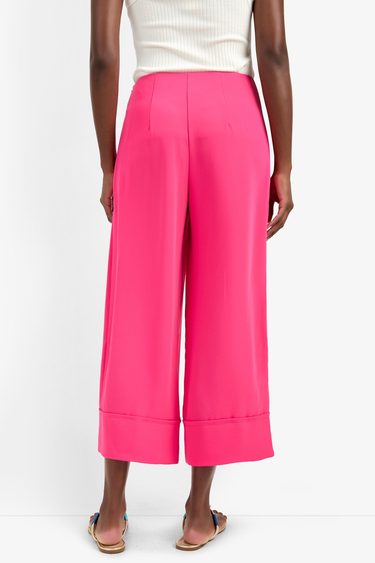 A person stands facing away in bright pink BAHOOKA PANT wide-leg, cropped pants (100% polyester) and a white sleeveless top with flat sandals, set against a plain white background.