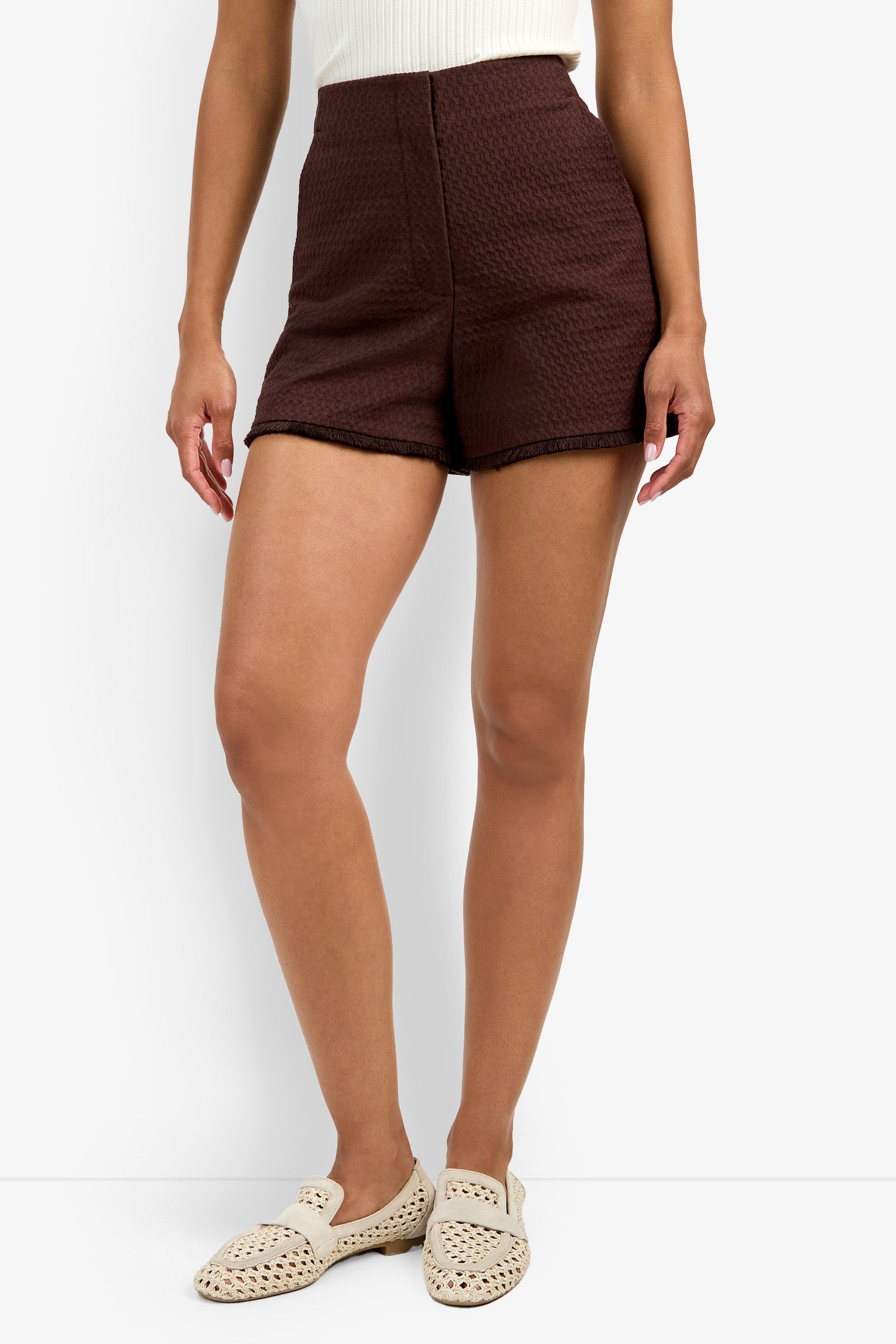 Someone is wearing the Valera Fringe Short in a textured dark brown style, paired with a white sleeveless top and cream woven slip-on shoes, standing against a plain white background.