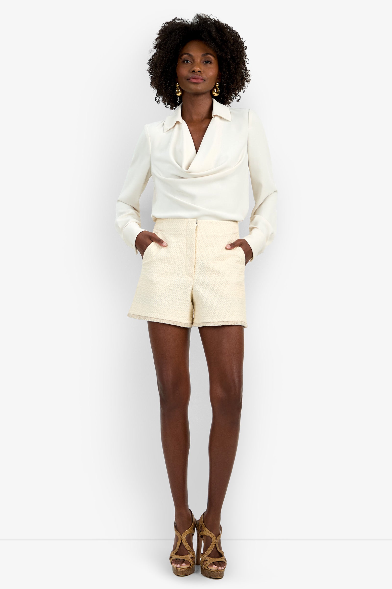 A woman stands with her hands in her pockets against a plain white background, wearing cream textured Valera Fringe Shorts, a white long-sleeve blouse, and brown high-heeled sandals.