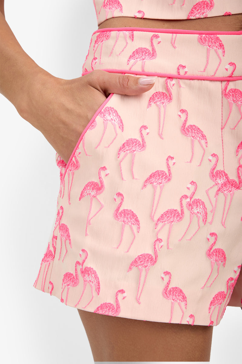 Wearing the Piped Jilene Short in light pink with a pink flamingo pattern, a person stands with one hand in the pocket, showing only their lower torso and upper thigh.