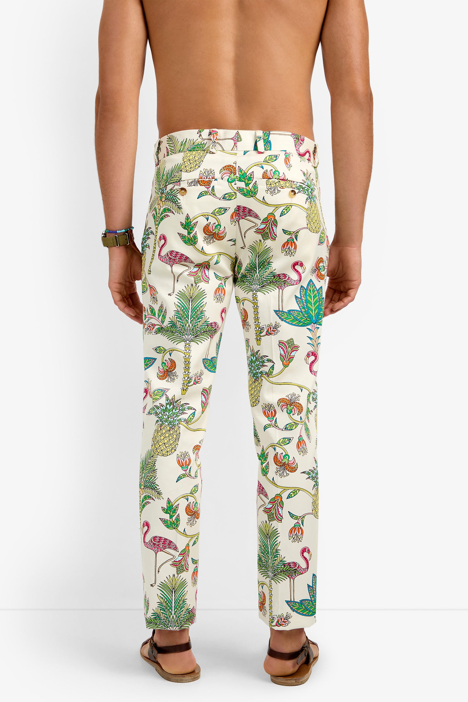 A person stands barefoot, facing away from the camera, wearing CLYDE SLIM TROUSER in a colorful tropical print with flamingos, plants, and birds; only their lower back and legs are visible.