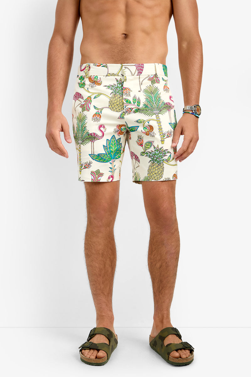 A man wears the LAWRENCE SHORT—patterned swim shorts crafted from stretch fabric with tropical flamingo, pineapple, and green leaf prints—paired with sandals and a wristwatch, set against a plain white background.