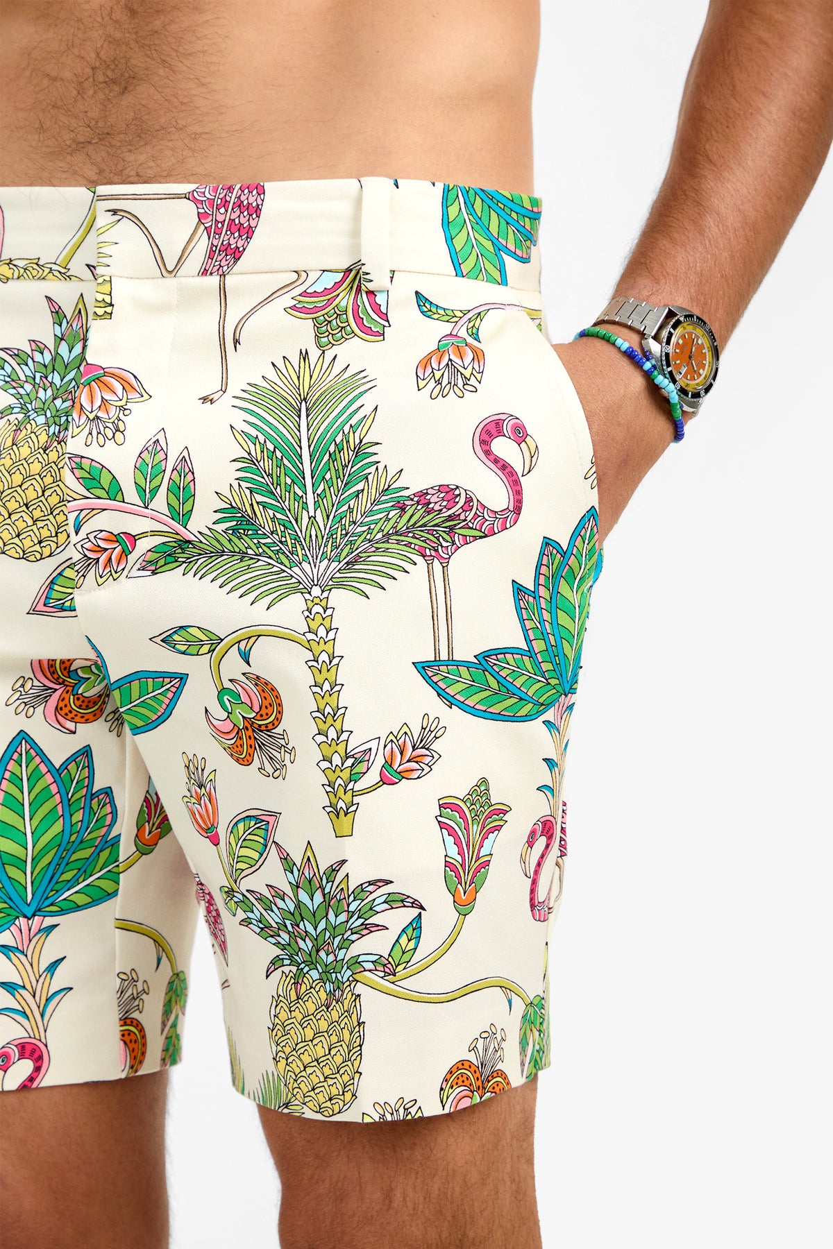 Someone wears the LAWRENCE SHORT, a comfortable cotton-blend short with colorful tropical prints of flamingos, pineapples, and palm trees; one hand is in their pocket and a silver wristwatch is visible.