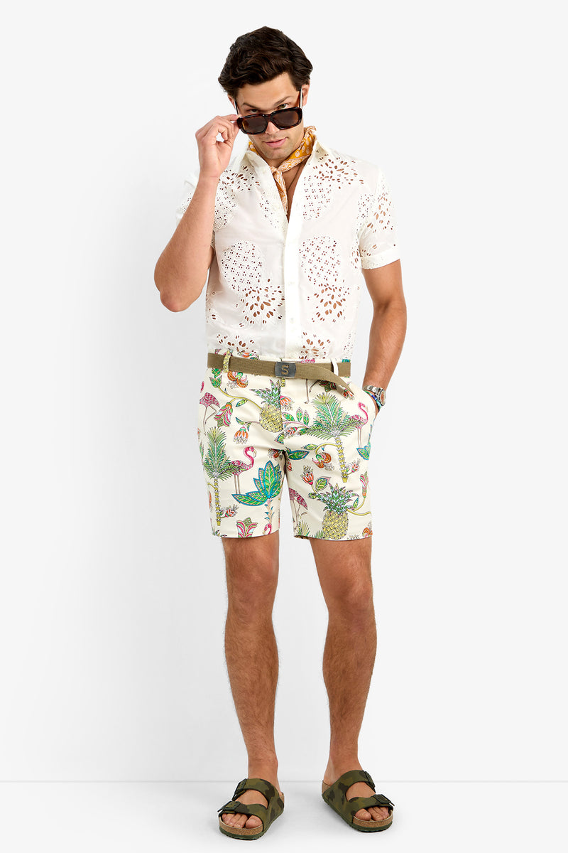 A man stands against a white background wearing sunglasses, a patterned scarf, a white cotton lycra eyelet shirt, the LAWRENCE SHORT in tropical print, olive green sandals, and a brown belt.
