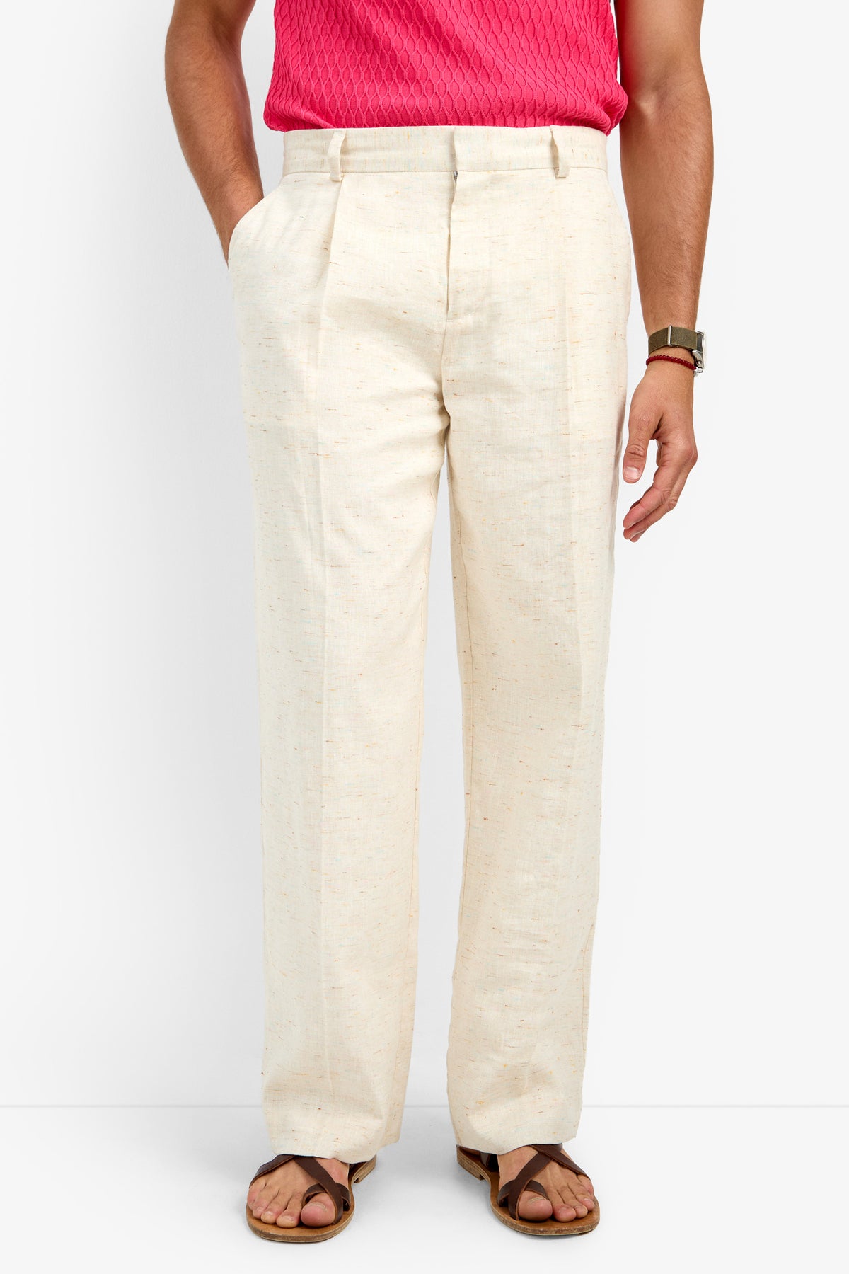 A person stands against a plain white background wearing the CLYDE WIDE LEG TROUSER in cream, with hands in pockets, styled with a pink textured shirt, sandals, and a watch.