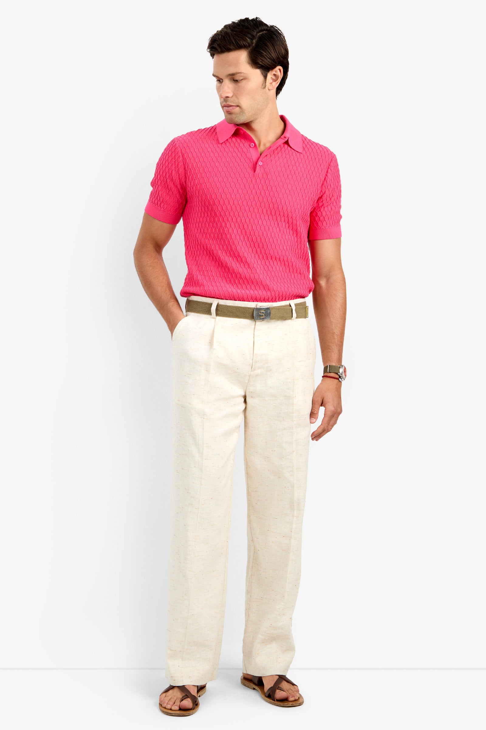 A man stands against a plain background, wearing a bright pink cotton lycra polo shirt, CLYDE WIDE LEG TROUSER in Linen Affair, a brown belt, watch, and brown sandals. He looks down with his hands in his pockets.