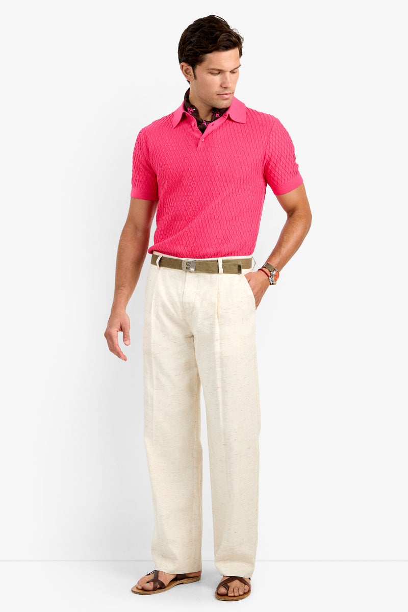 A man stands against a white background wearing the CLYDE WIDE LEG TROUSER in beige with pleats, paired with a pink short-sleeve knit polo, brown belt, and brown sandals. He looks down to his left with one hand in his pocket.