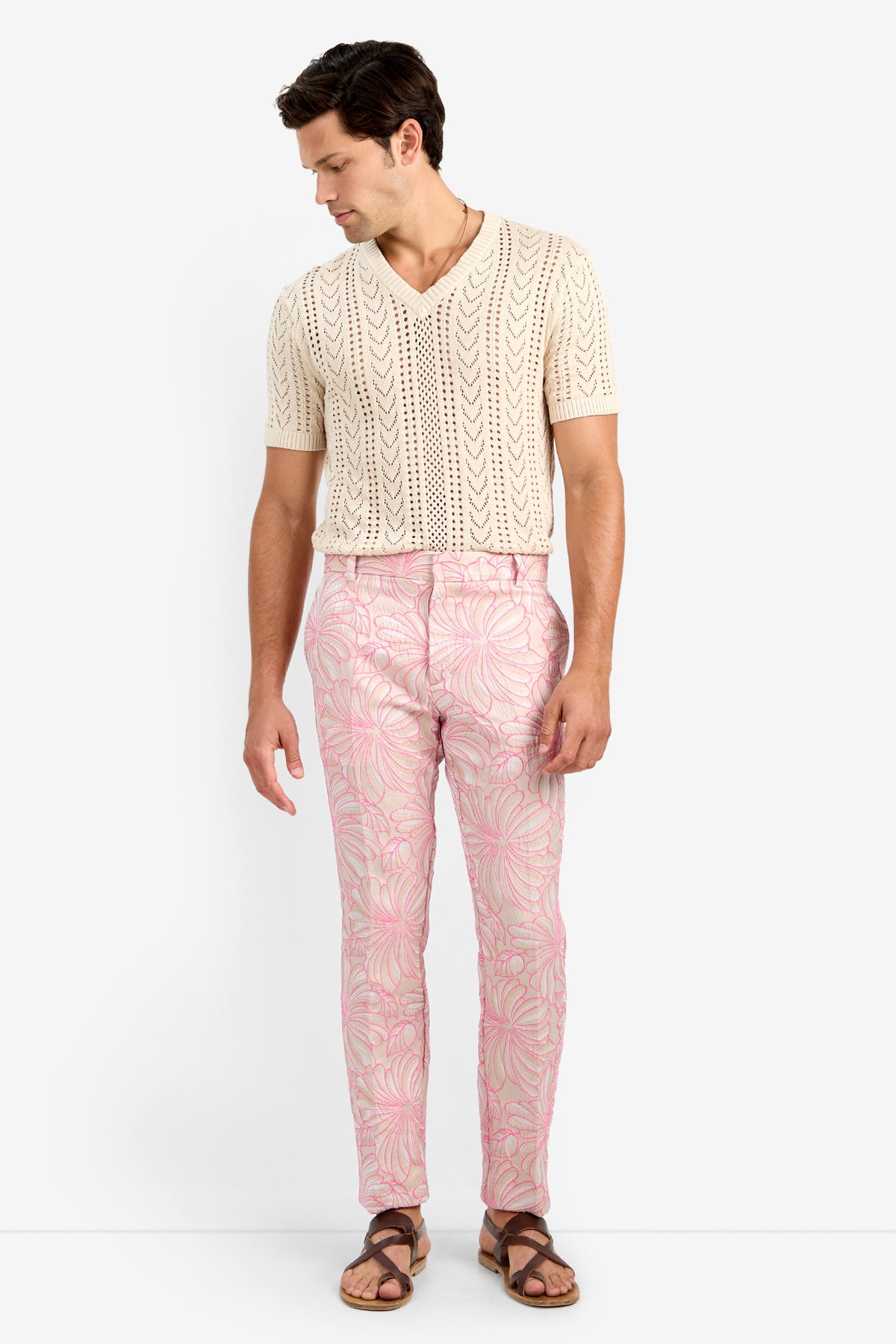 A man stands against a white background, wearing pink leafy-patterned CLYDE SLIM TROUSER made from lycra, a cream cotton short-sleeve knit shirt, and brown sandals. He looks down to his left with arms relaxed at his sides.