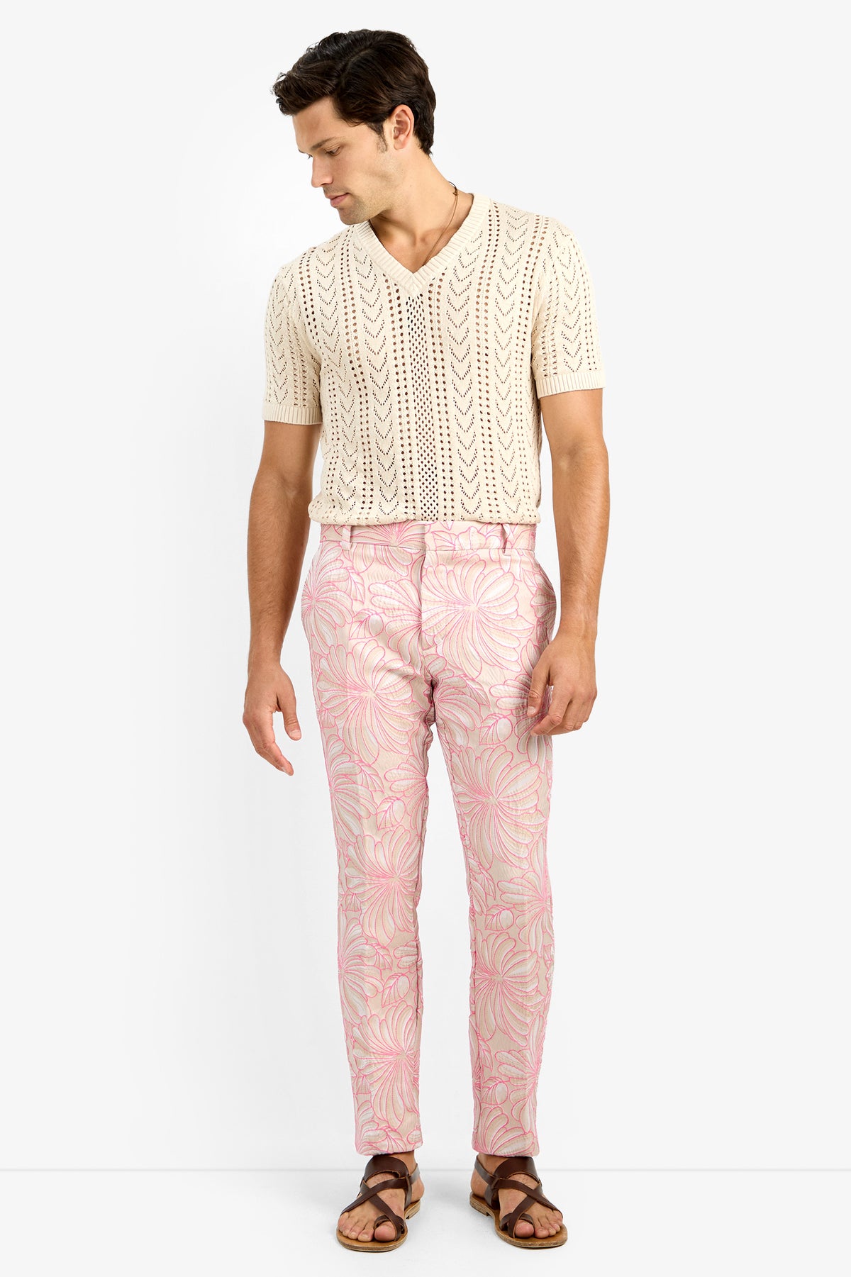A man stands against a white background wearing the Keanu Crochet V-neck Sweater in cream, paired with pink patterned pants and brown sandals. He looks down and to the side, arms relaxed at his sides.