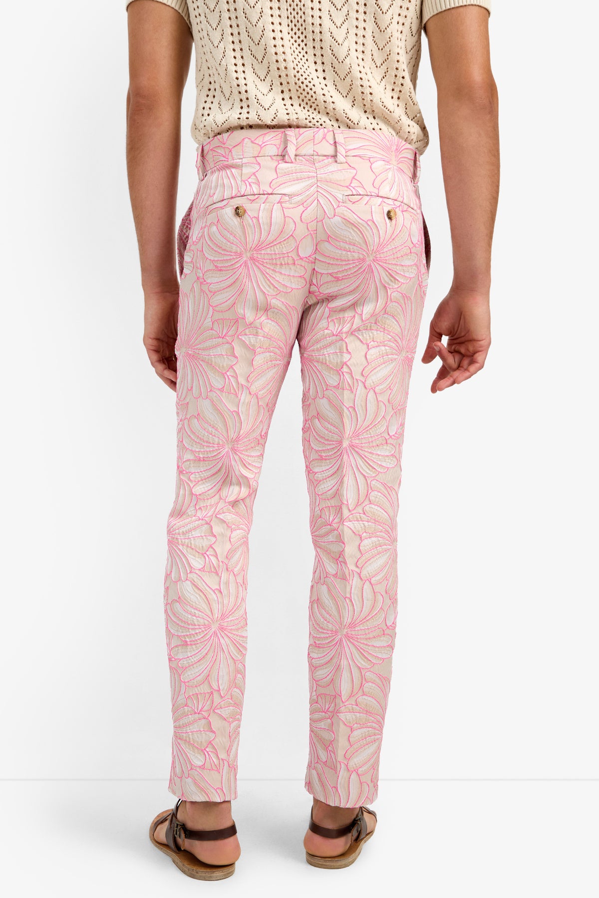 A person wearing CLYDE SLIM TROUSER in light pink with a large floral pattern, a beige knit short-sleeve shirt, and brown sandals, shown from the back against a plain white background.