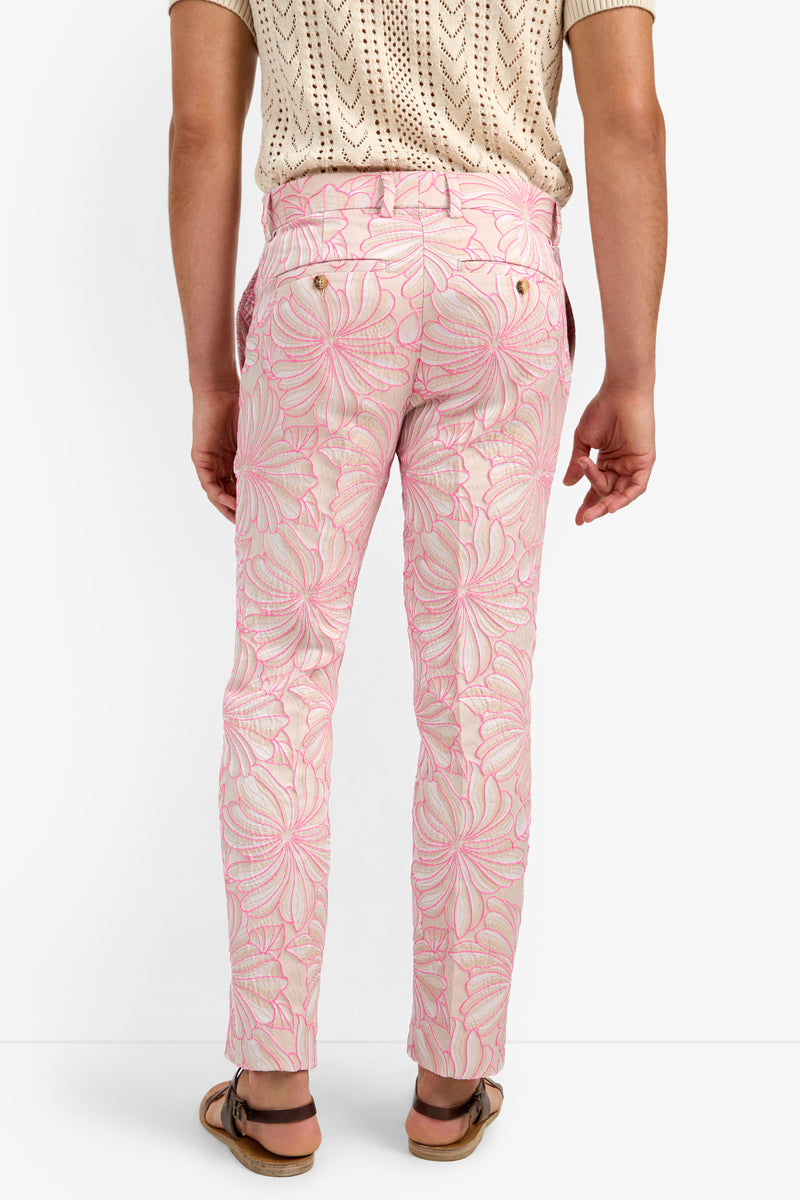 A person wearing CLYDE SLIM TROUSER in light pink with a large floral pattern, a beige knit short-sleeve shirt, and brown sandals, shown from the back against a plain white background.