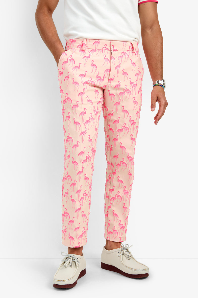A person wearing CLYDE SLIM TROUSER in Flamingos (light pink with a flamingo pattern), a white shirt with pink trim, a silver watch, and beige shoes with brown soles, standing against a plain white background.