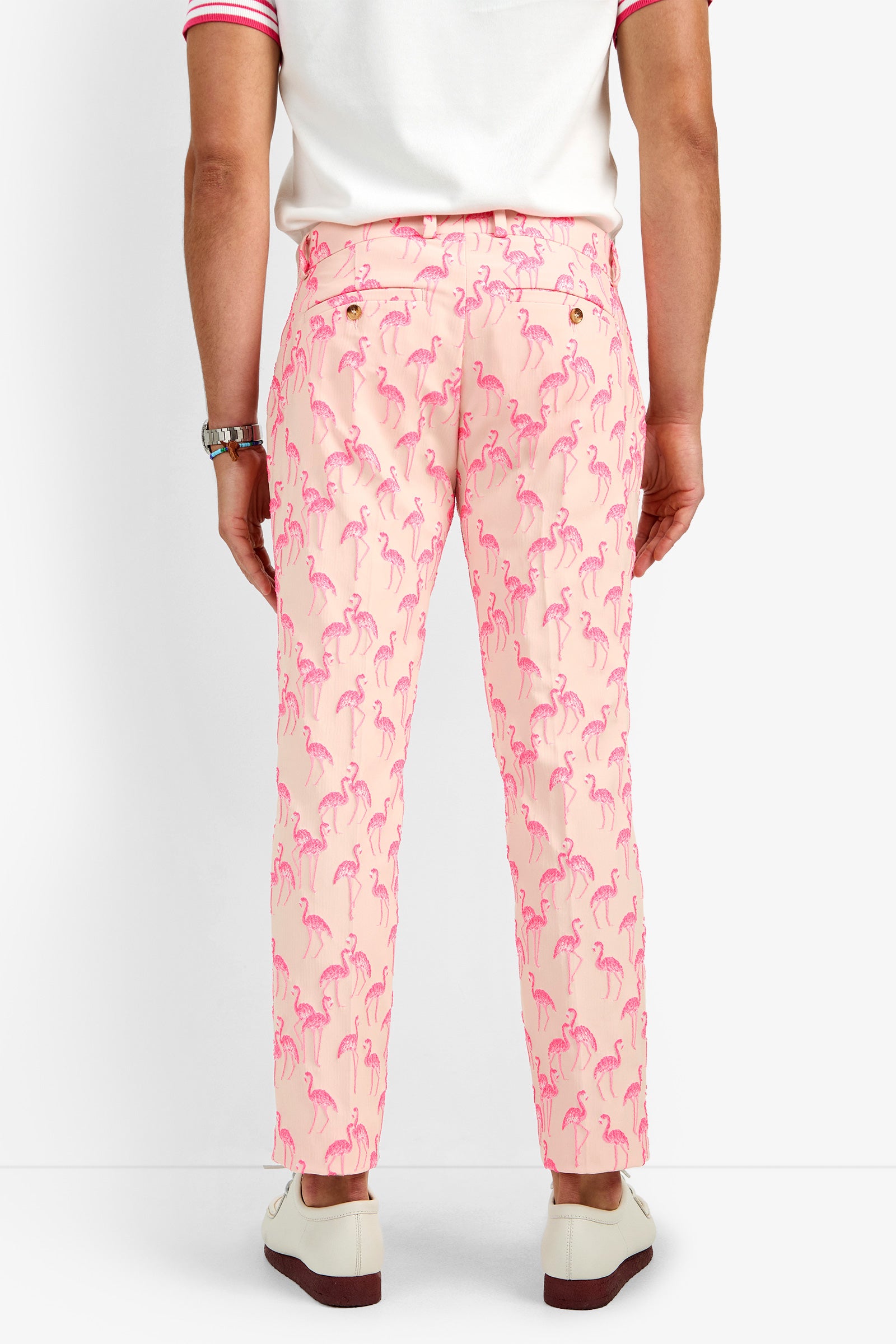 A person faces away from the camera, dressed in CLYDE SLIM TROUSER—light pink pants with bright pink flamingo prints made of soft polyamide—paired with a white shirt featuring pink trim and white loafers.