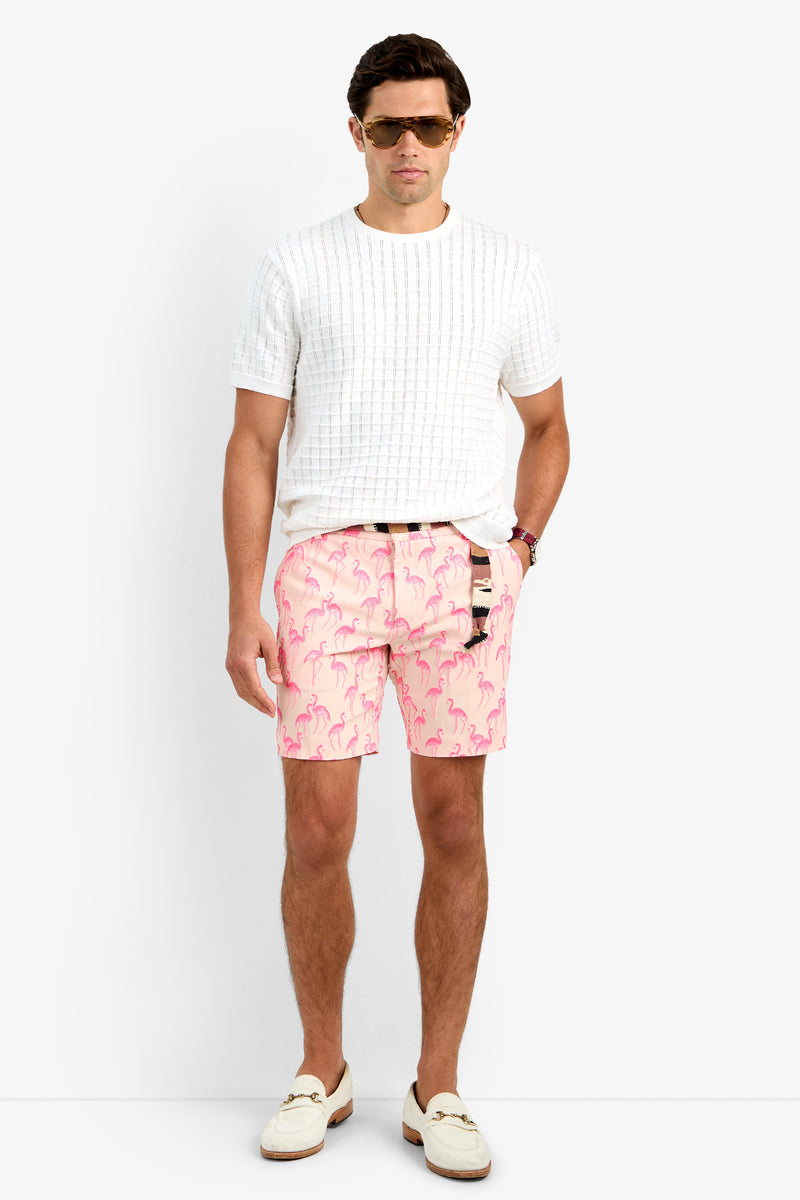 A man wearing sunglasses, a white textured polyamide short-sleeve shirt, LAWRENCE SHORT in Flamingos, a brown belt, and white loafers stands against a plain white background with one hand in his pocket.