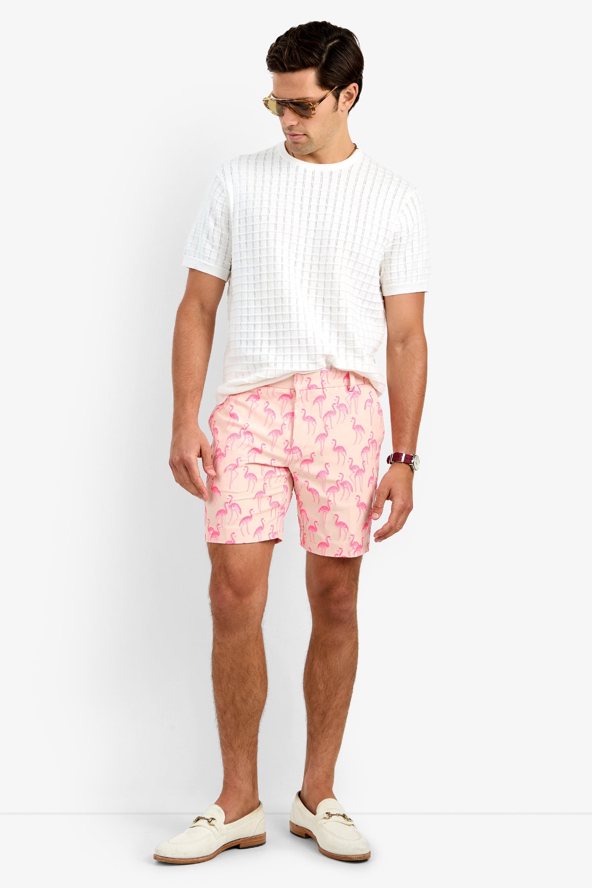 A man wearing sunglasses, a textured white polyamide short-sleeve shirt, LAWRENCE SHORT pink flamingo-patterned shorts, a watch, and white loafers stands against a plain white background, looking down slightly.