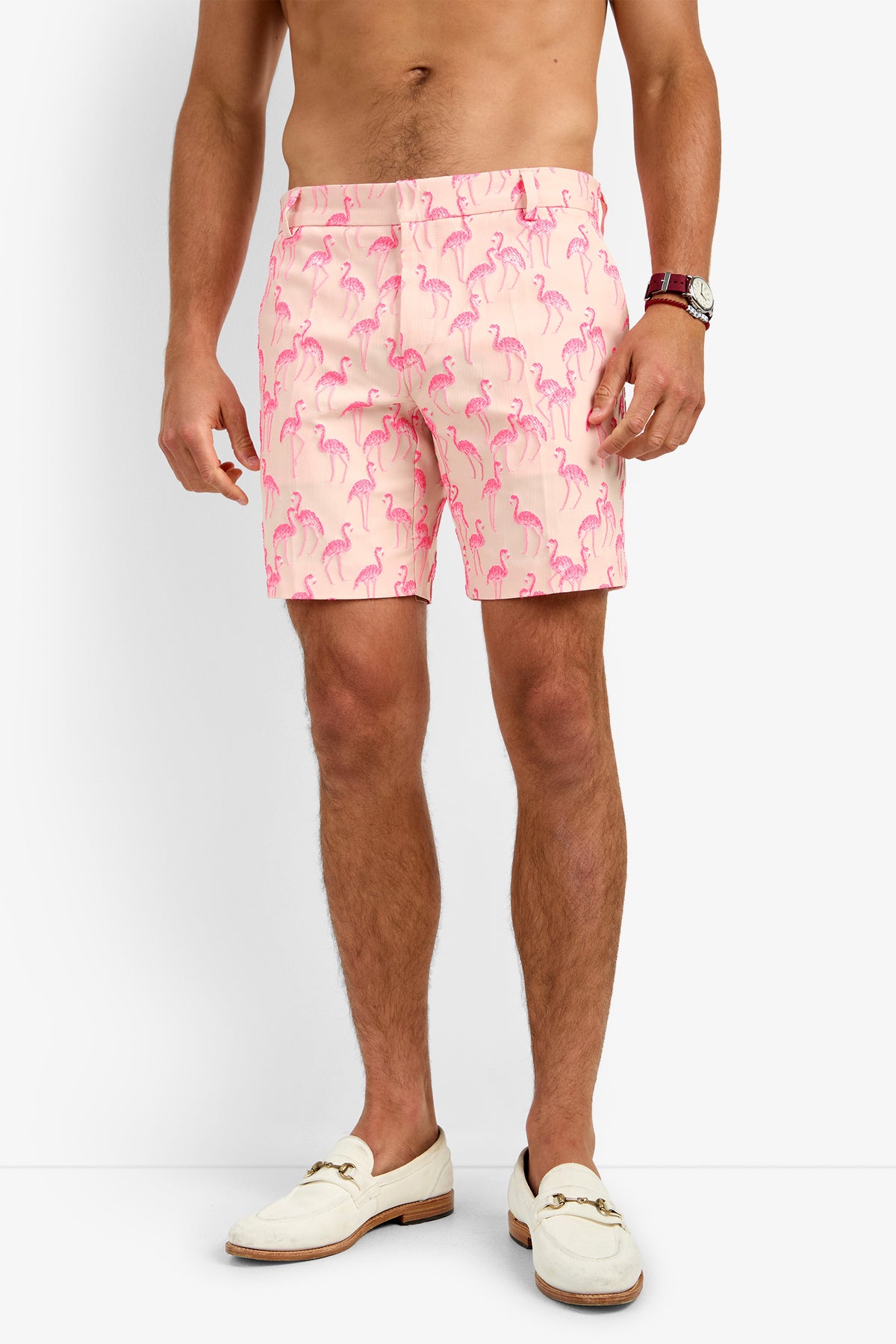 A man wears light pink LAWRENCE SHORTS with a pink flamingo print, a brown watch, and cream loafers. He is shirtless and standing against a white background.