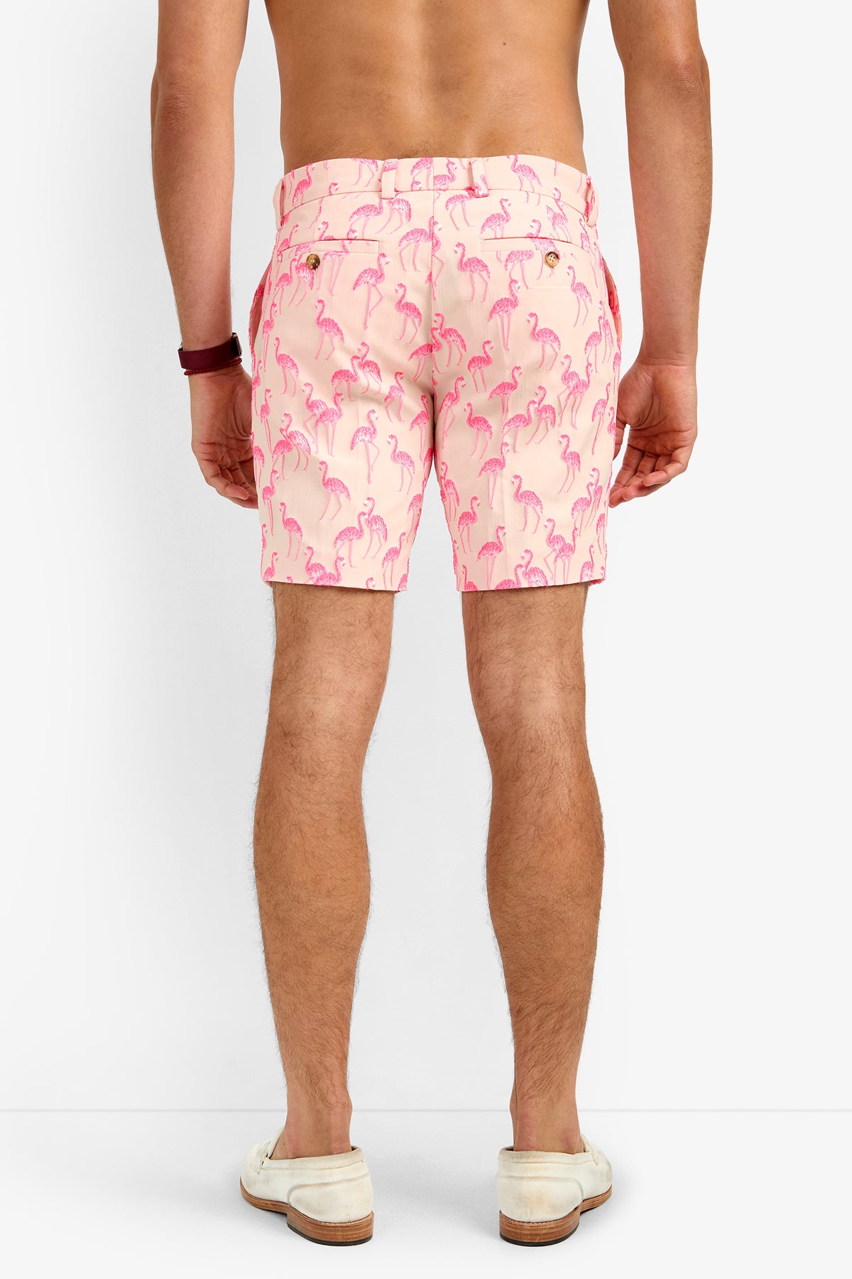 A man stands barefoot and shirtless, facing away, wearing LAWRENCE SHORT—light pink polyester shorts featuring a repeating pink flamingo pattern.