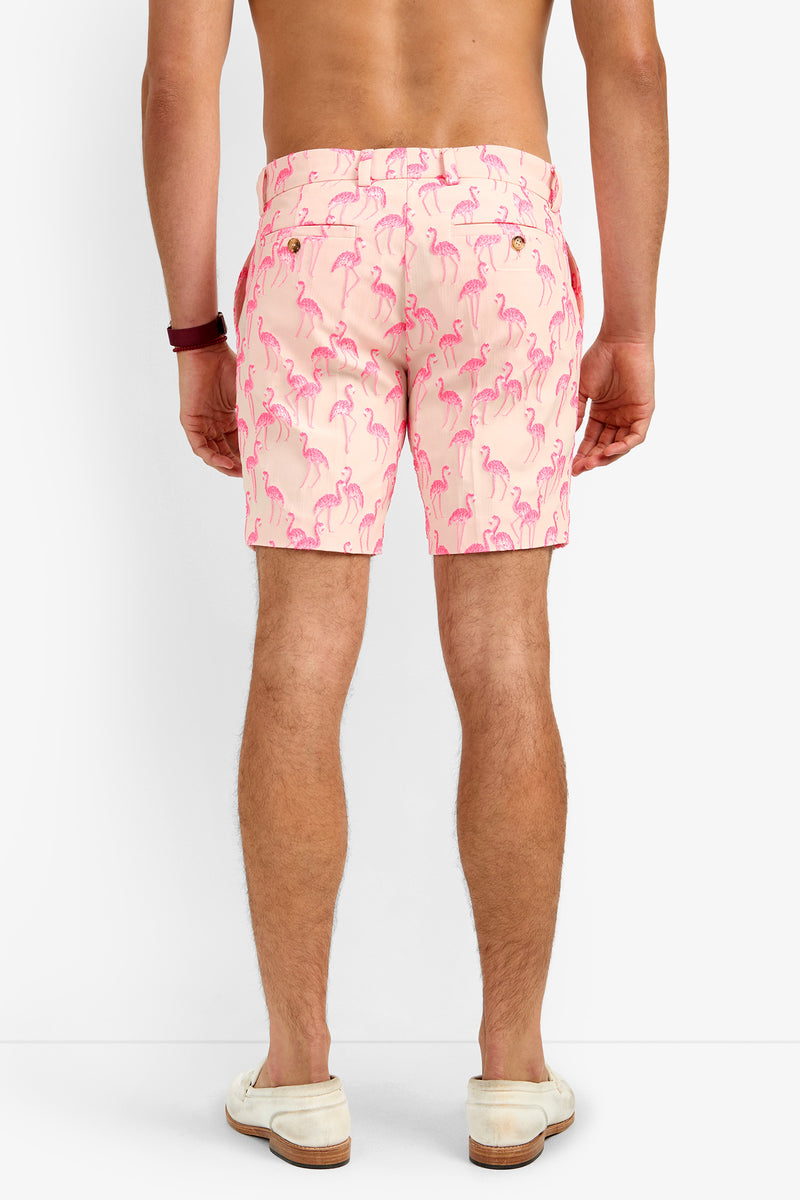 A man stands barefoot and shirtless, facing away, wearing LAWRENCE SHORT—light pink polyester shorts featuring a repeating pink flamingo pattern.