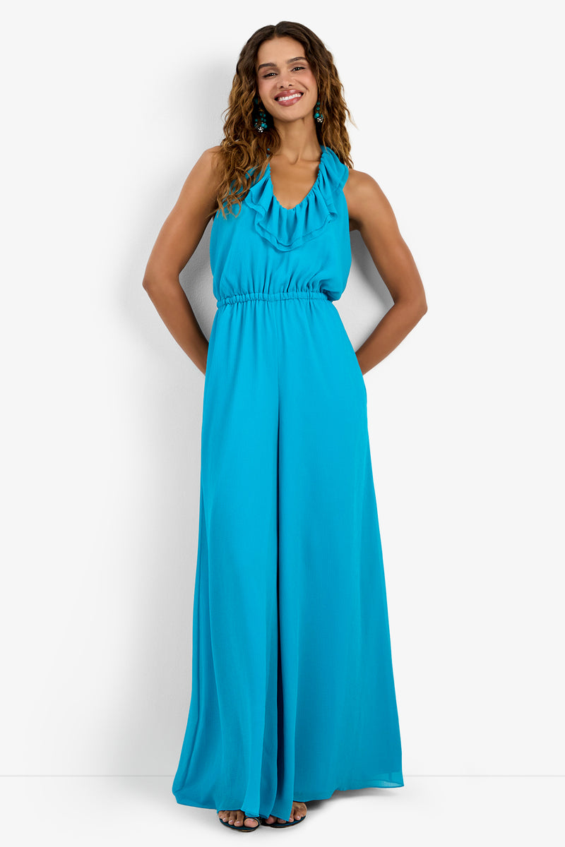 A woman smiles with hands in pockets, wearing the JACKSONVILLE JUMPSUIT—a sleeveless, floor-length, bright turquoise halter-neck jumpsuit in 100% polyester with a ruffled neckline and wide legs. She poses against a white background.