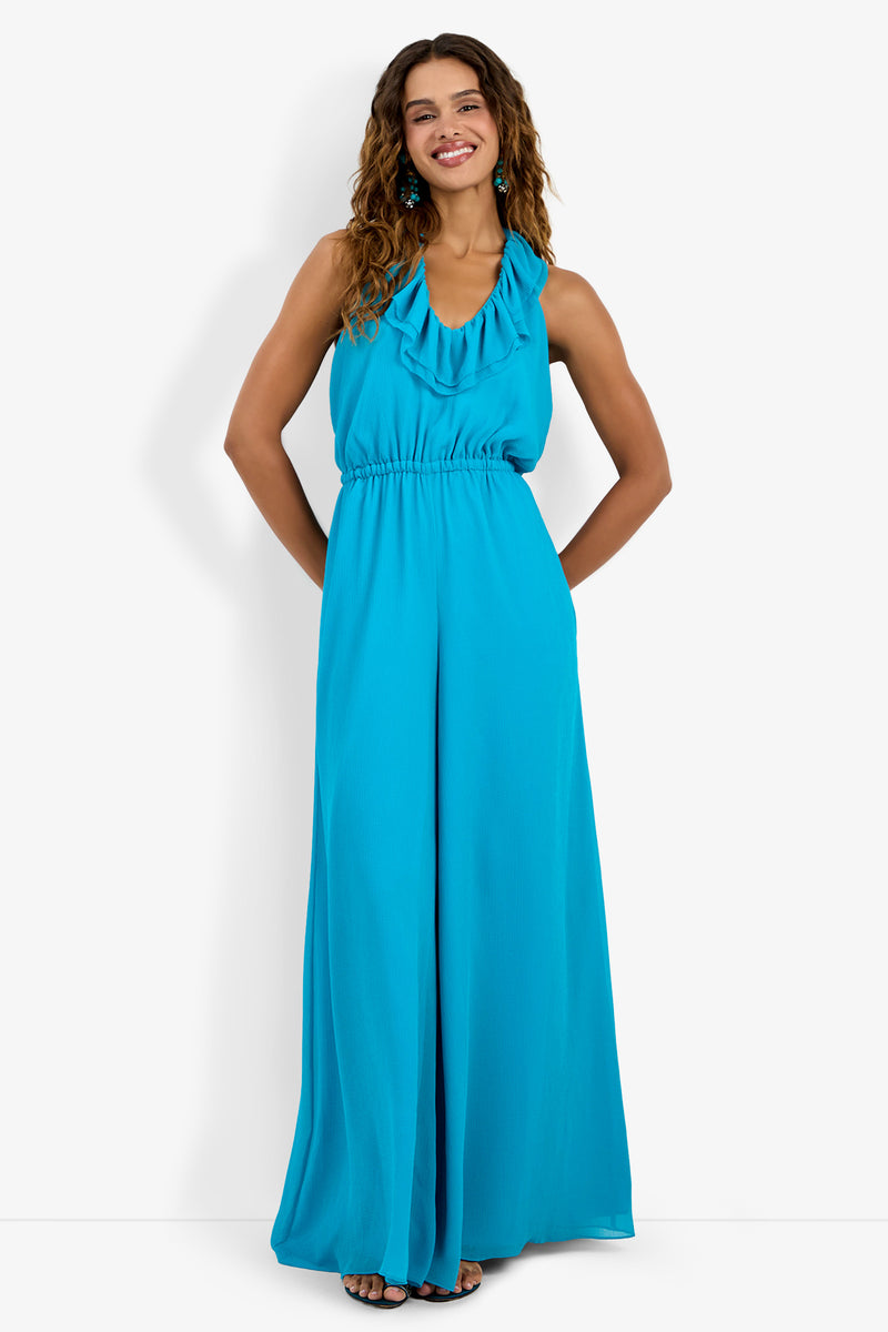 A woman with curly hair smiles, hands in her pockets, wearing the Jacksonville Jumpsuit—a sleeveless, floor-length, bright blue halter jumpsuit with a ruffled neckline and gathered waist—against a plain white background.