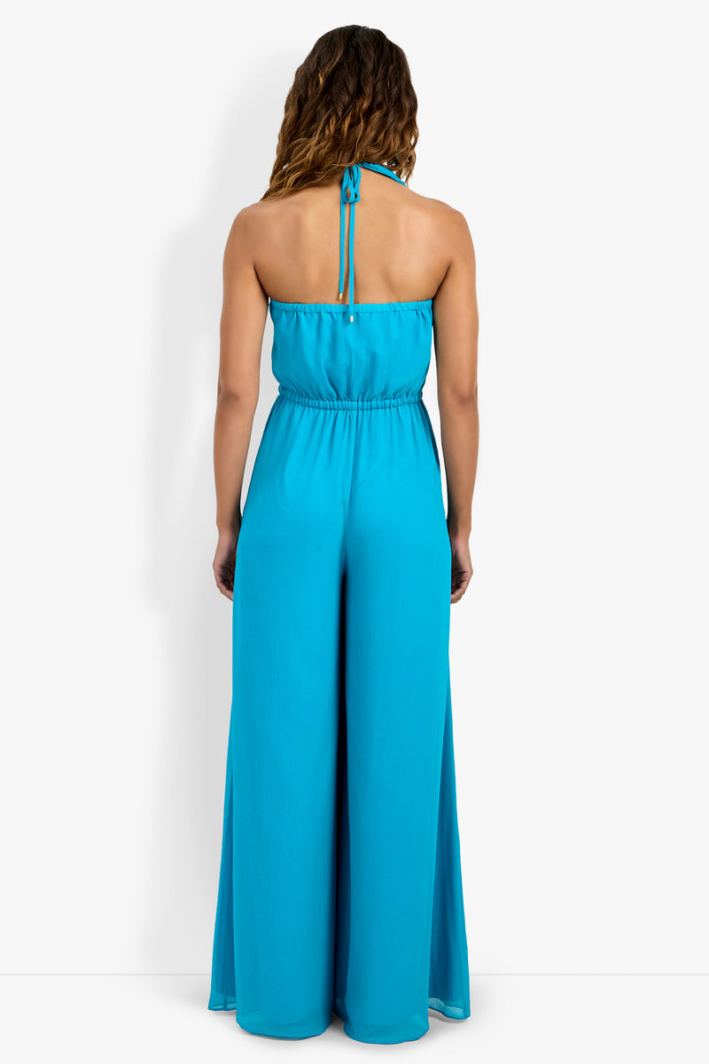 A woman faces away, wearing the Jacksonville Jumpsuit—a sleeveless, bright blue halter jumpsuit with wide-leg pants and an elastic waist. Her wavy brown hair is down, set against a plain white background.