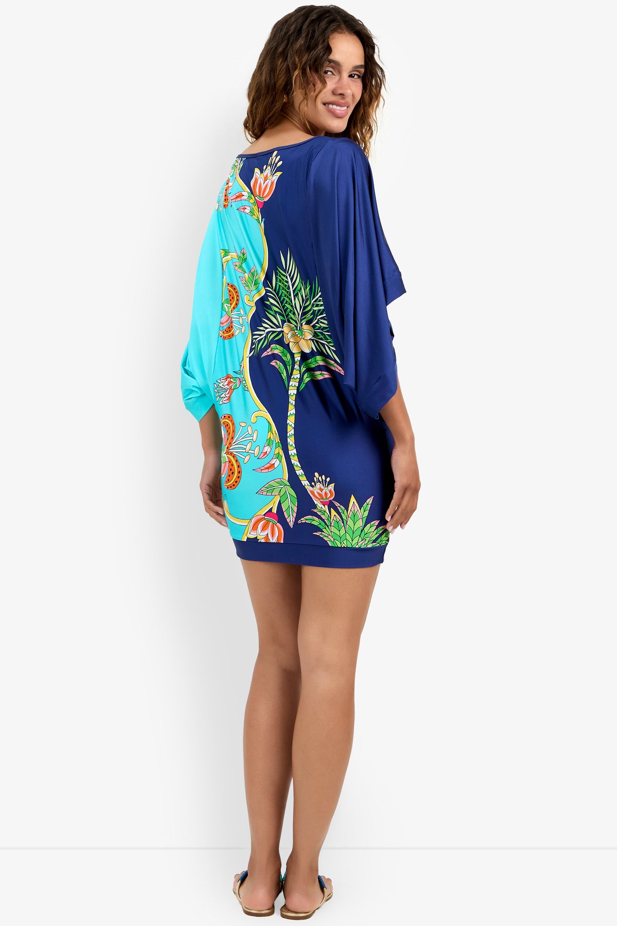 Woman smiling and standing in the Casablanca Dress, a short, loose-fitting dress featuring colorful tropical patterns with palm trees and flowers in blue, green, orange, and yellow. She glances over her shoulder.