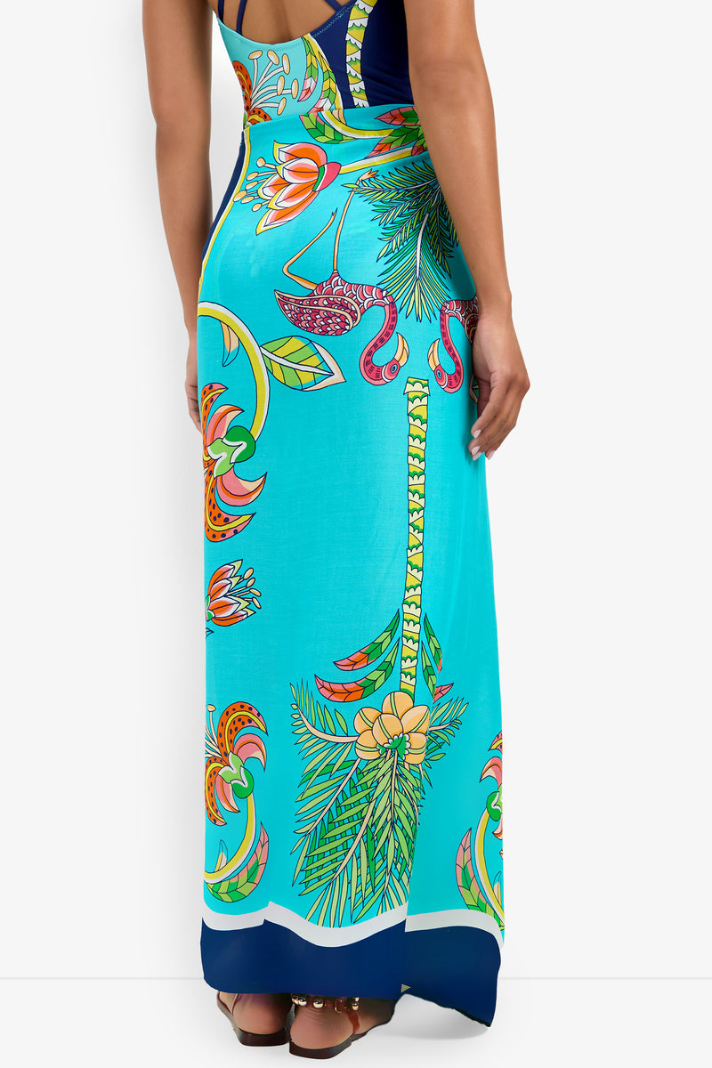 Someone wearing the Pareo, a long turquoise dress featuring vibrant tropical floral and palm leaf patterns, with a fitted waist and ankle-length flow.