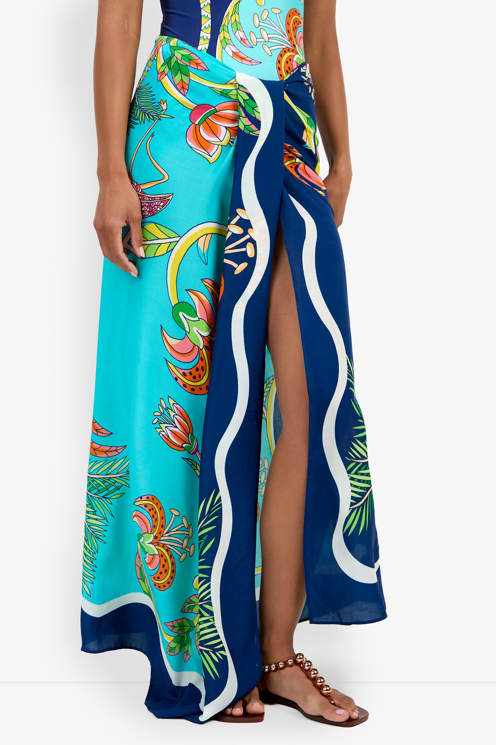 A person wearing a turquoise and blue floral-patterned Pareo with a high slit, styled with a dark top and beaded sandals.