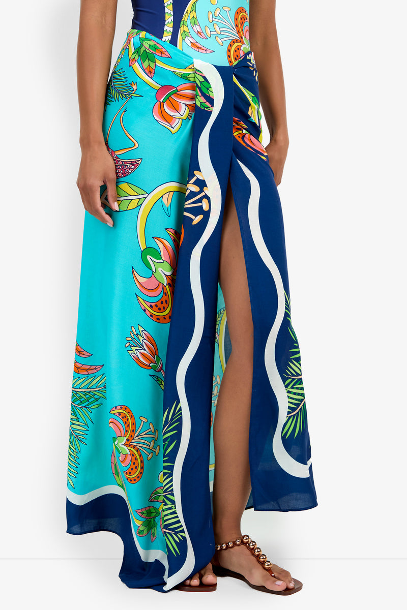 A person wearing a turquoise and blue floral-patterned Pareo with a high slit, styled with a dark top and beaded sandals.