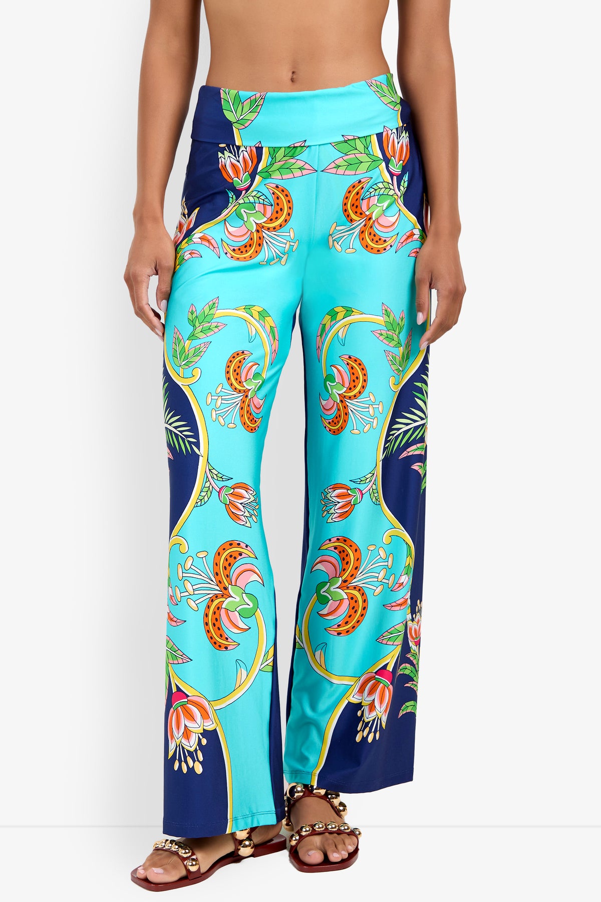 A woman models the Swim Pant—high-waisted, wide-leg turquoise pants with a vibrant floral and leaf print—paired with brown beaded sandals, shown from midriff to feet against a white background.