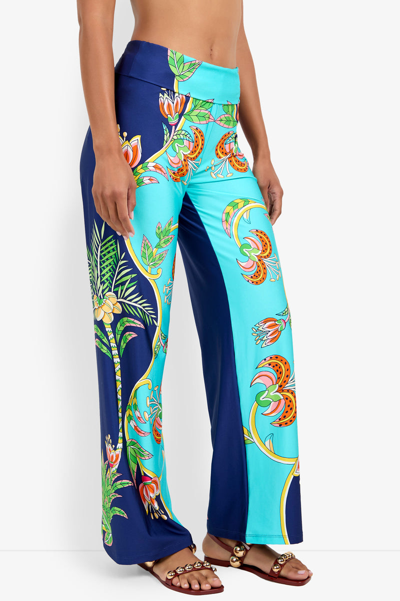 A person wears the Swim Pant, featuring bright blue and turquoise tropical floral and palm leaf prints, paired with brown sandals adorned with gold embellishments.