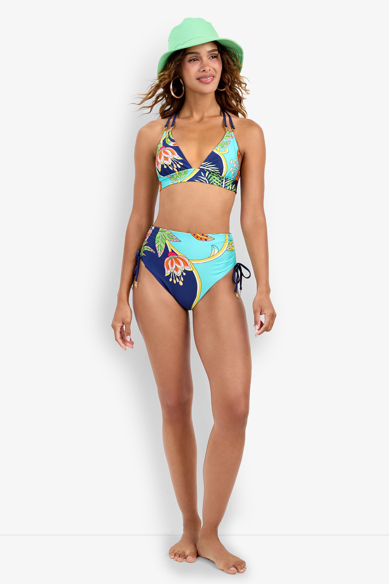 A woman smiles while wearing the Banded Halter Bikini Top and high-waisted floral bottoms, paired with a green bucket hat and large hoop earrings, standing barefoot against a plain white background.