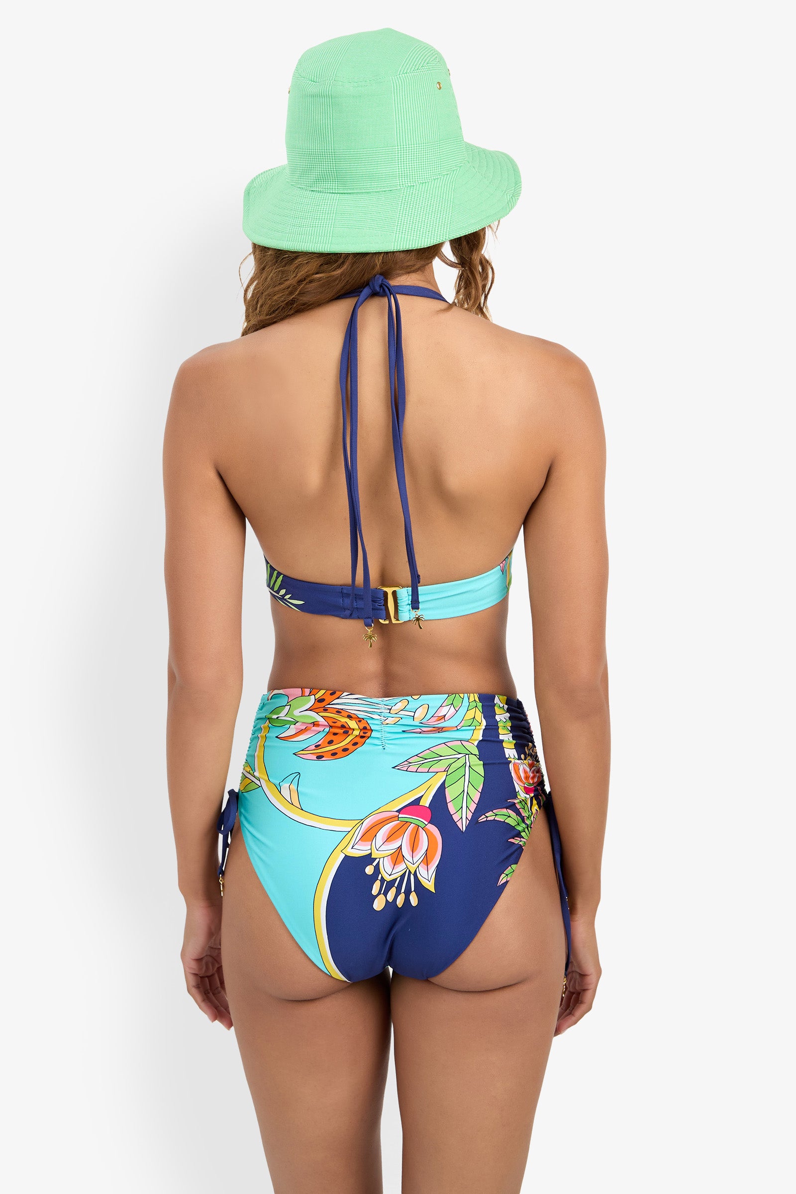 Woman faces away, wearing a green bucket hat and the Banded Halter Bikini Top—a blue, high-waisted floral bikini with a halter neck tie—standing before a plain white background.