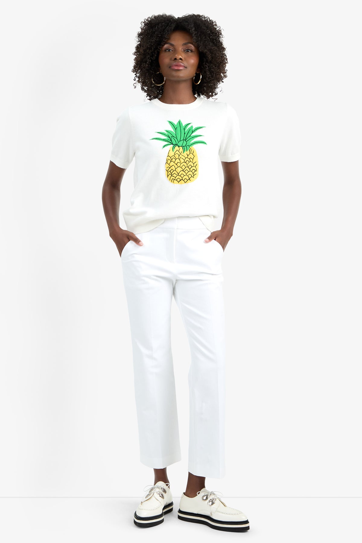 A woman with curly hair stands against a white background, hands in her pockets, wearing the MAUI OLD FASHIONED SWEATER, white pants, and white shoes with black soles.